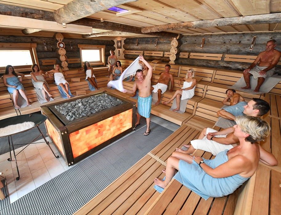 Menschen entspannen in einer großen Sauna mit Holzbänken und einem zentralen Ofen.
