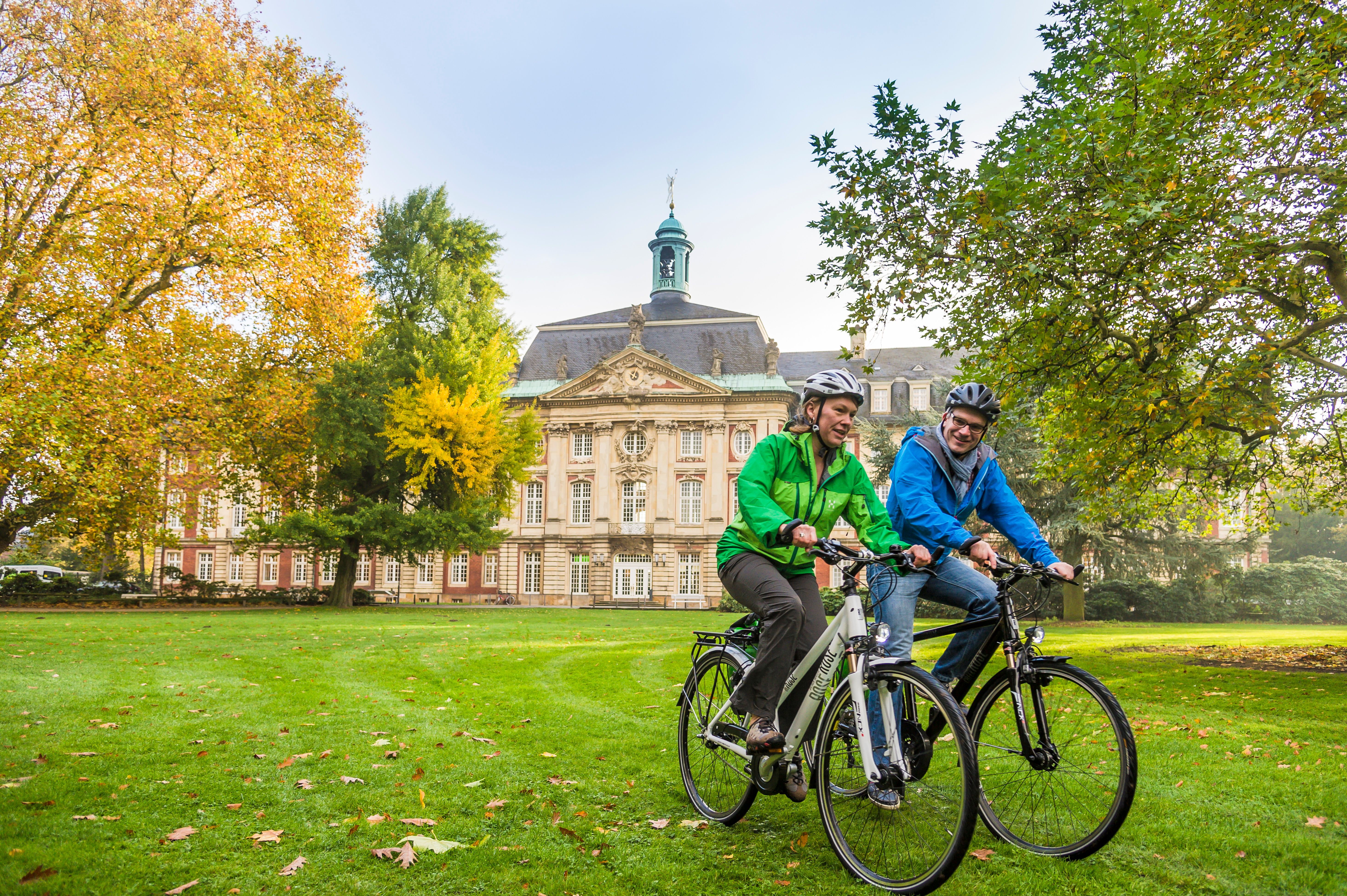 Zwei Personen fahren mit Fahrrädern vor dem Schloss Münster auf einer grünen Wiese.
