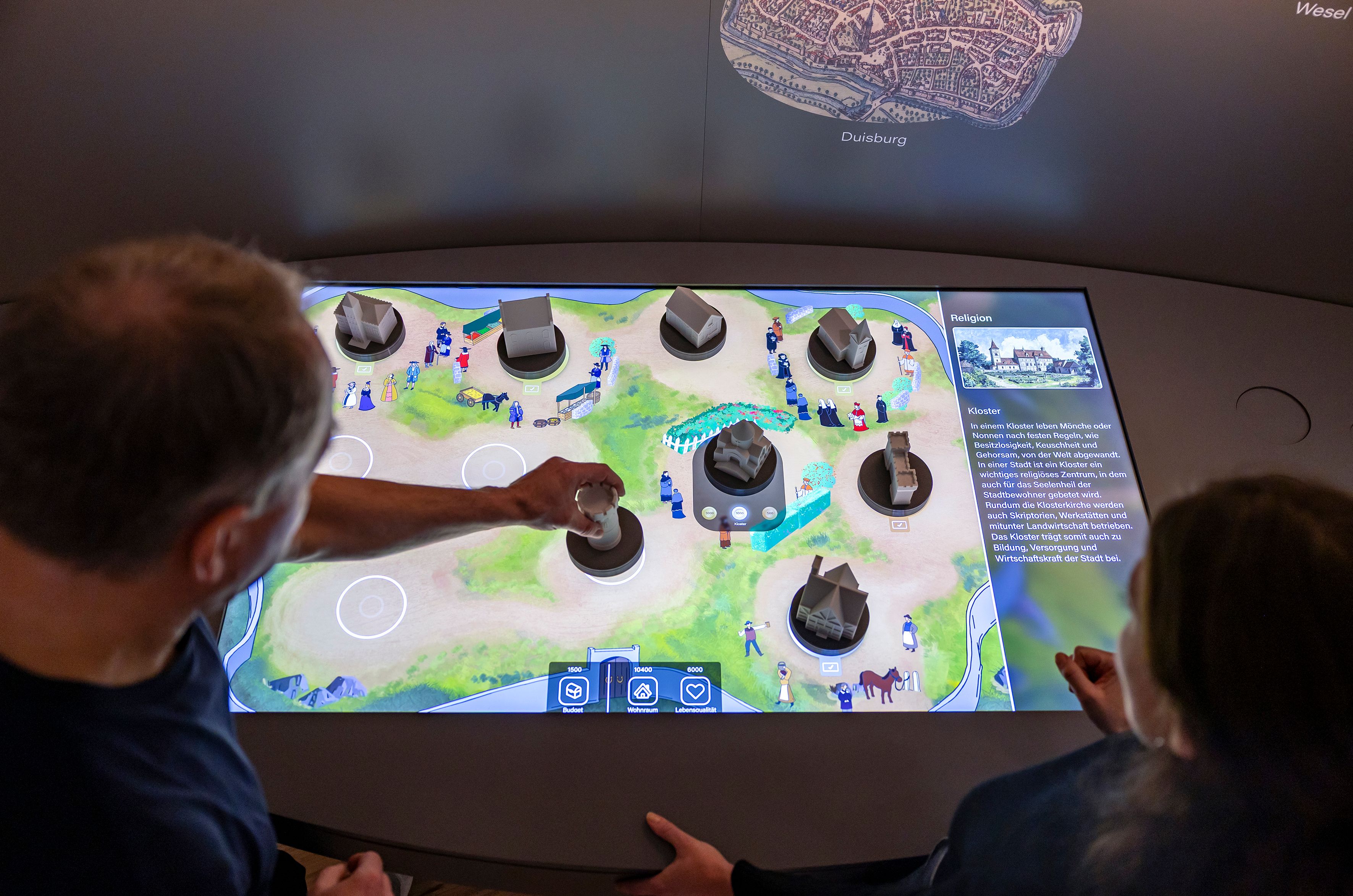 Zwei Personen interagieren mit einem interaktiven Bildschirm, der eine mittelalterliche Stadt darstellt.
