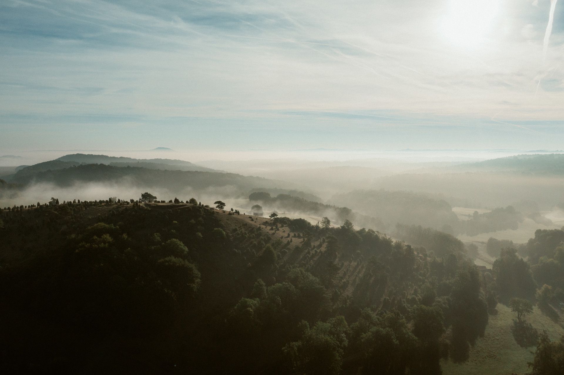 Morgennebel über dem Kalvarienberg Alendorf, sanfte Hügel und Bäume im diffusen Licht der aufgehenden Sonne.
