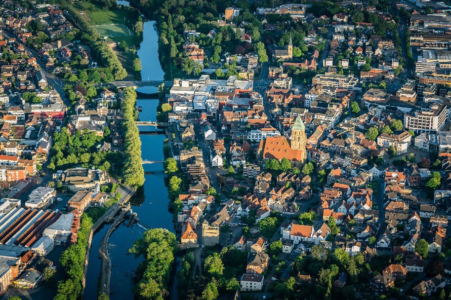 Luftaufnahme der Innenstadt von Rheine. Ein Fluss fließt durch die Stadt, umgeben von Gebäuden und einer Kirche.