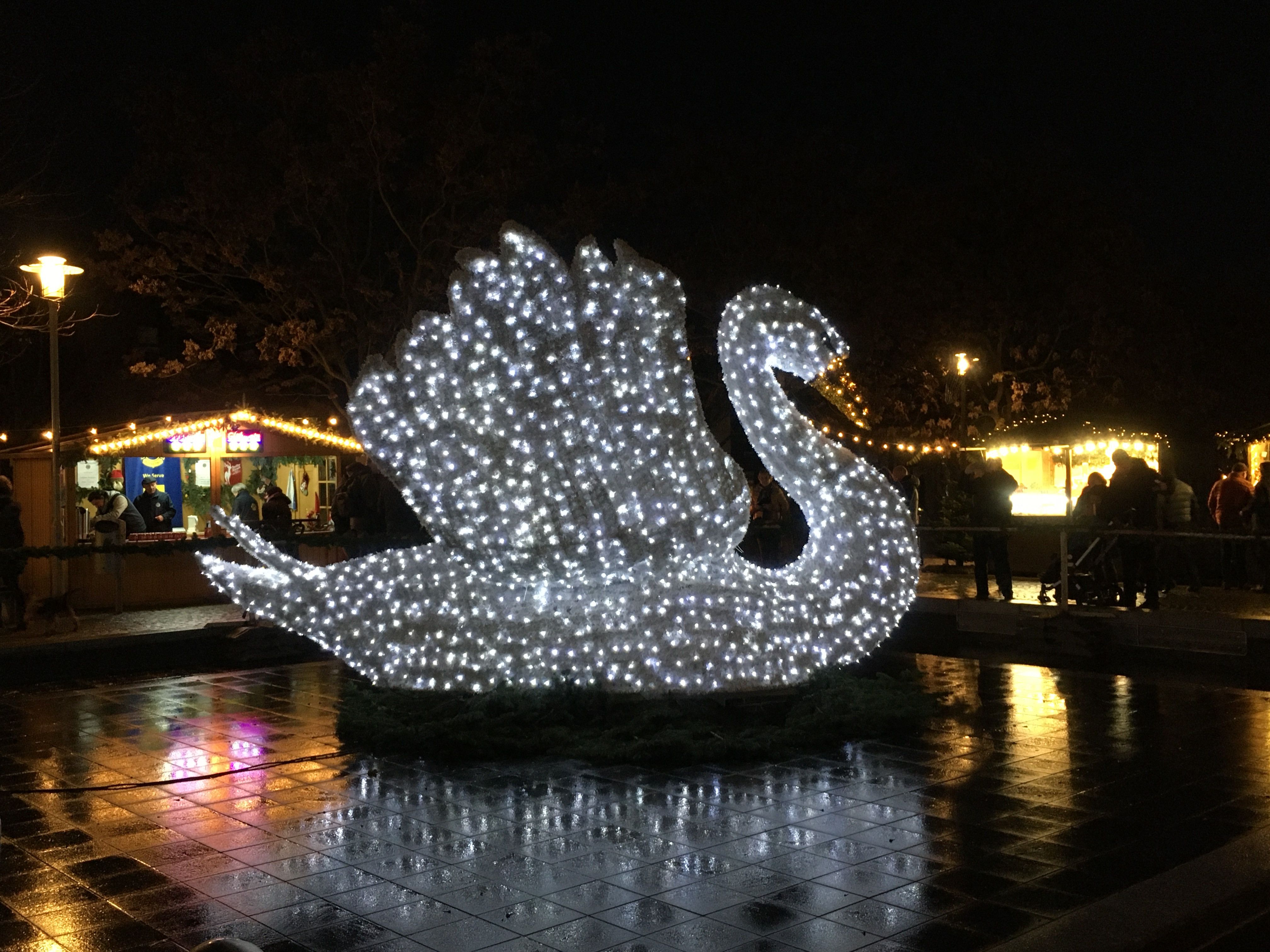 Leuchtende Schwanenfigur auf dem Klever Weihnachtsmarkt bei Nacht, umgeben von Buden und spiegelndem nassem Pflaster.