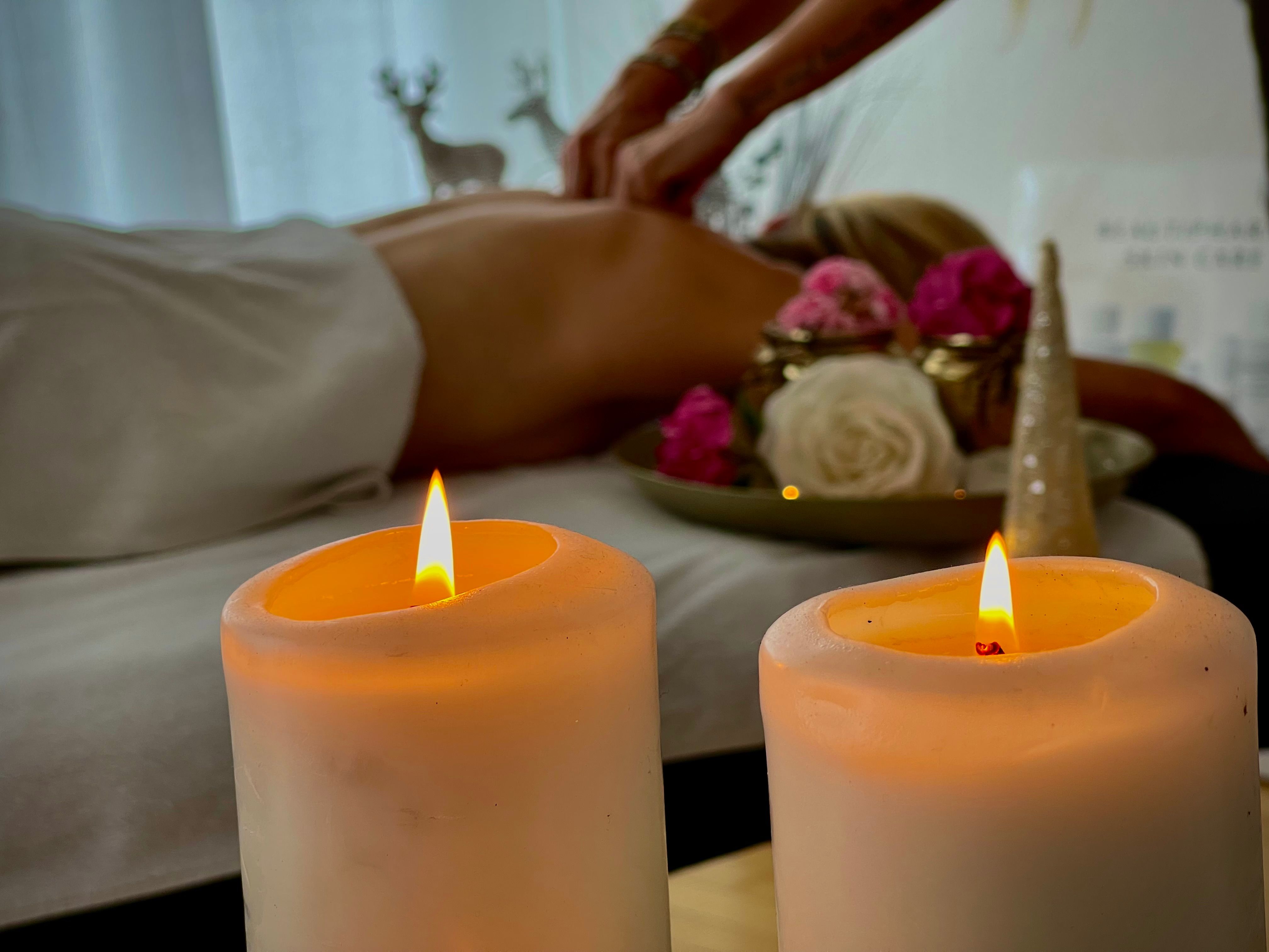 Zwei brennende Kerzen im Vordergrund, im Hintergrund eine Person, die eine Massage erhält.