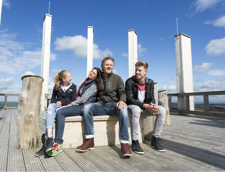 Vier Personen sitzen auf einer Bank auf einem Baumwipfelpfad mit blauem Himmel im Hintergrund.