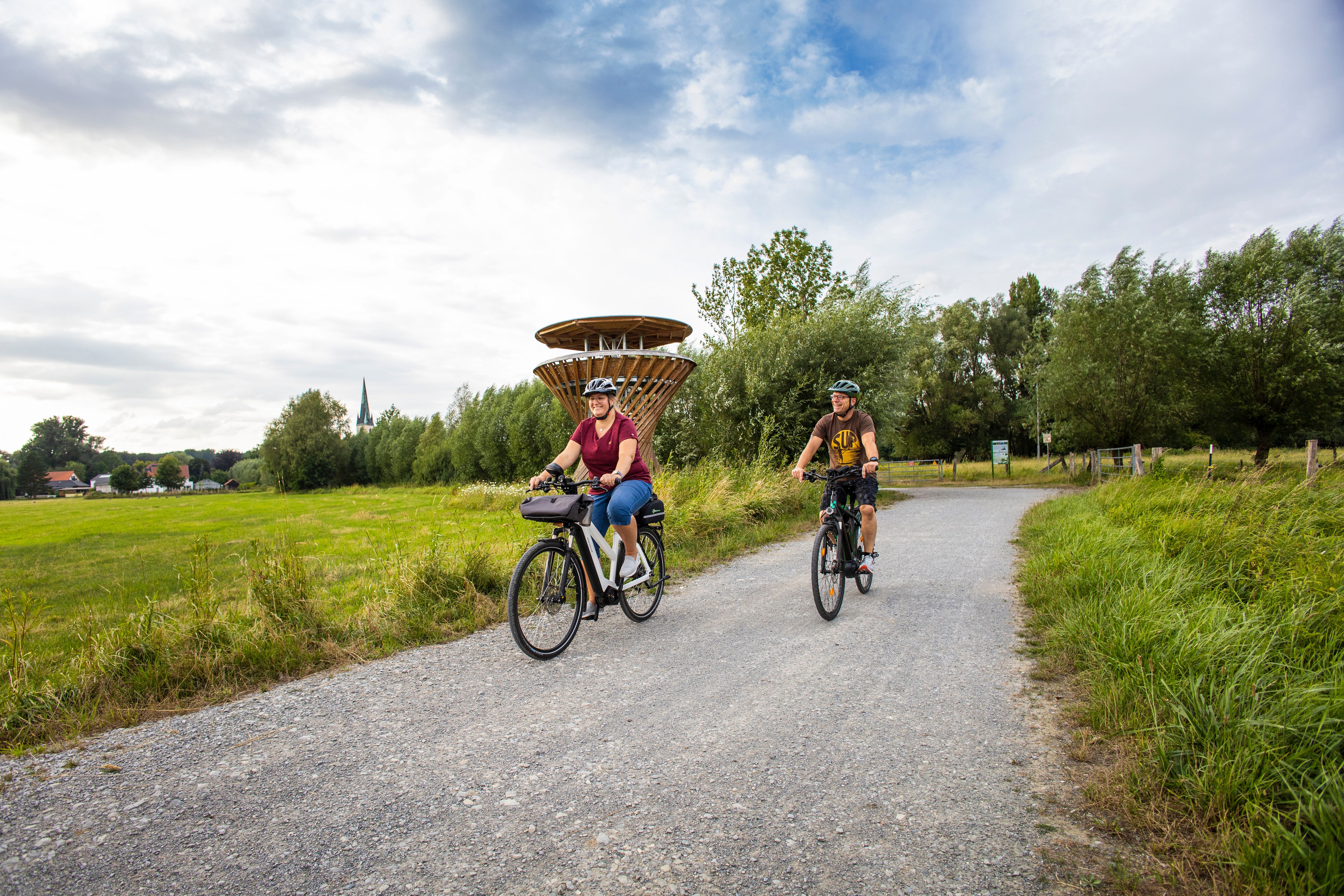 Zwei Radfahrer fahren entspannt auf einem Schotterweg umgeben von grüner Wiesenlandschaft an einem hölzernen Aussichtsturm vorbeifahren.