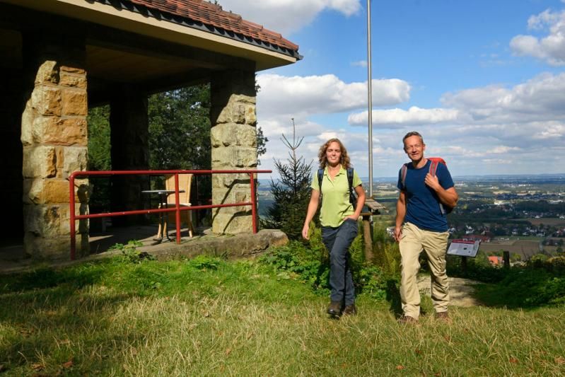 Zwei Personen wandern bei sonnigem Wetter an einem Steingebäude vorbei mit Blick auf eine weite Landschaft.