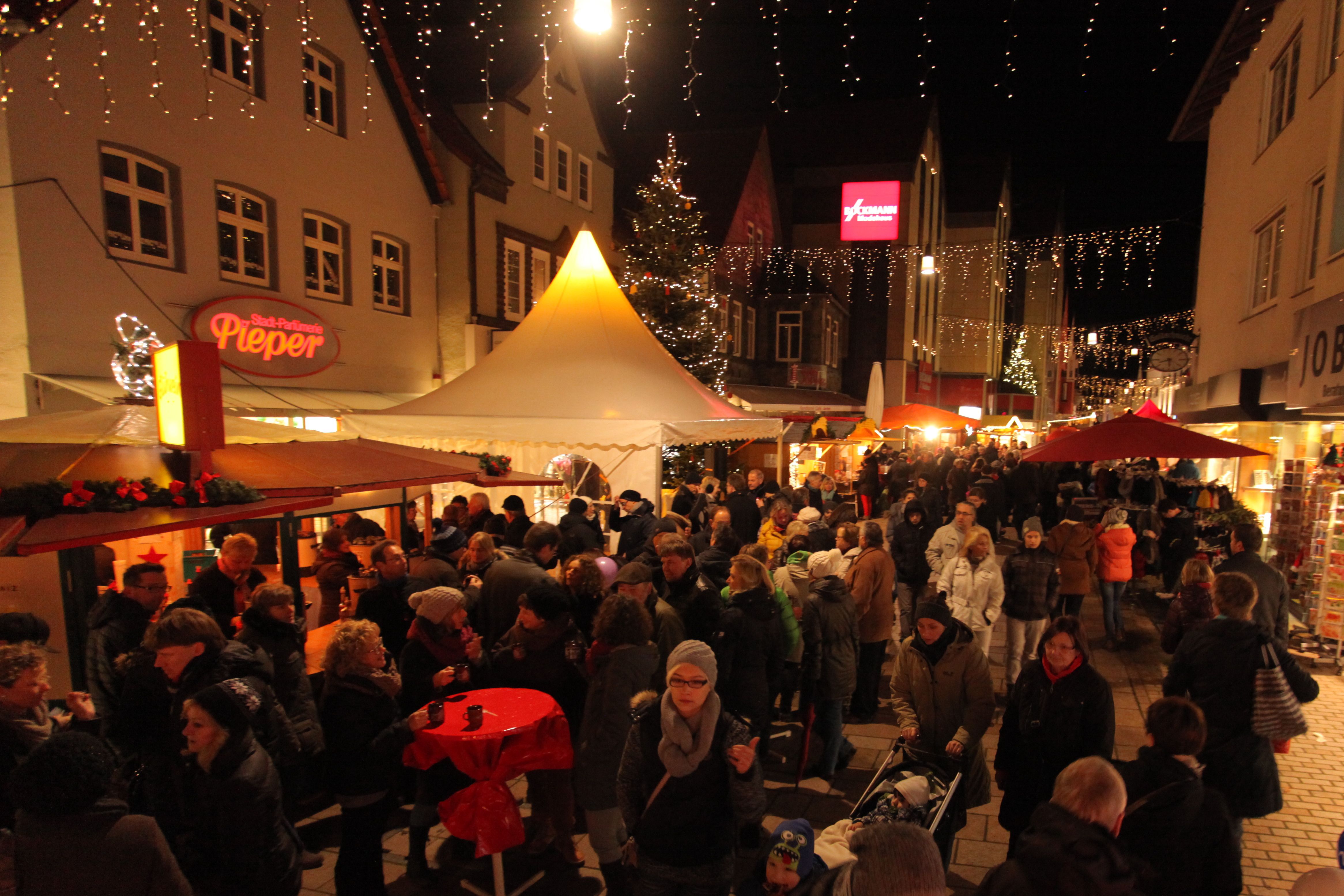Der Weihnachtsmarkt in Lübbecke ist gut besucht. Menschen schlendern zwischen festlich beleuchteten Ständen, während Lichterketten die Straße schmücken.