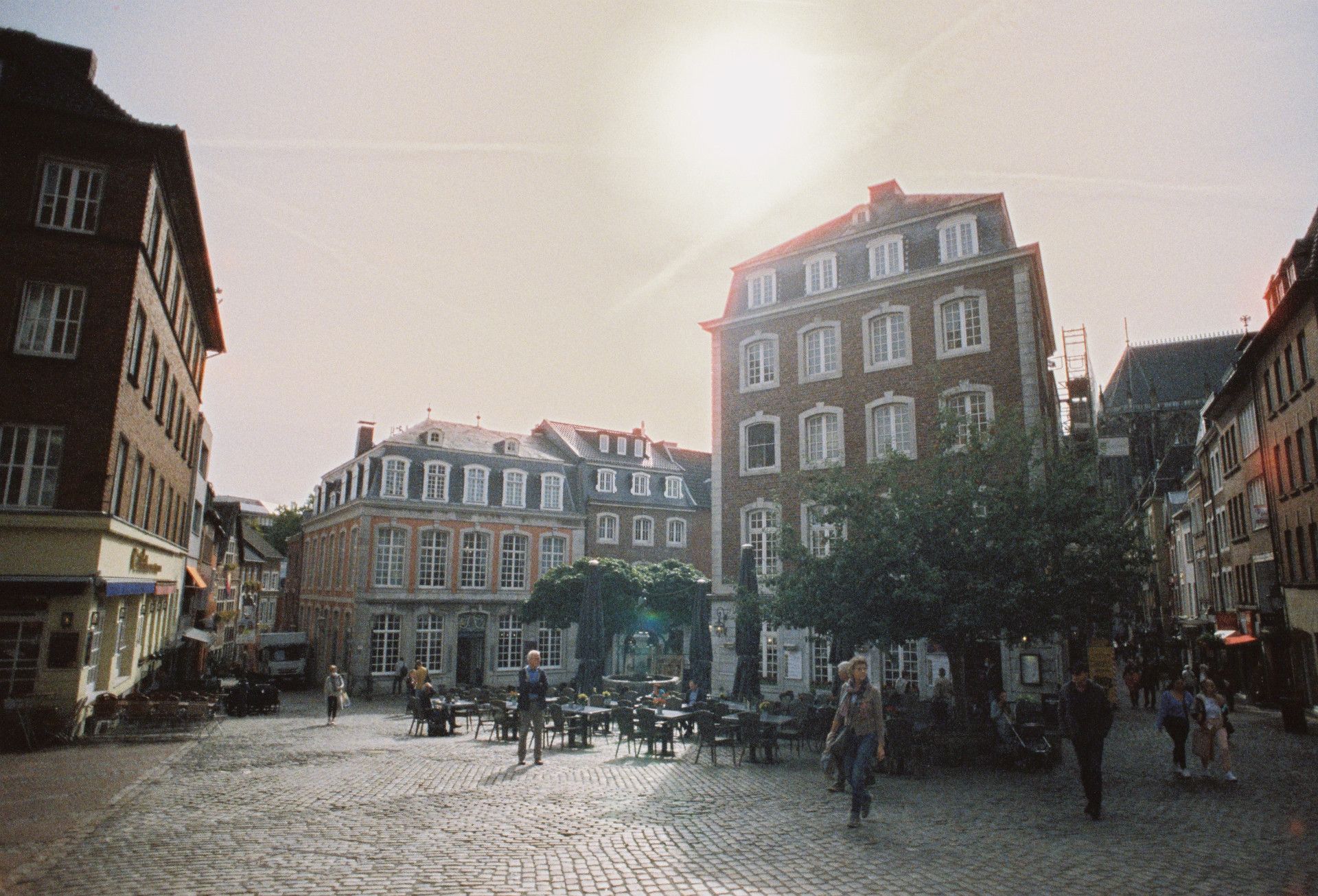 Sonnenbeschienener Platz in der Aachener Altstadt mit historischen Gebäuden und Menschen, die spazieren gehen oder sitzen.