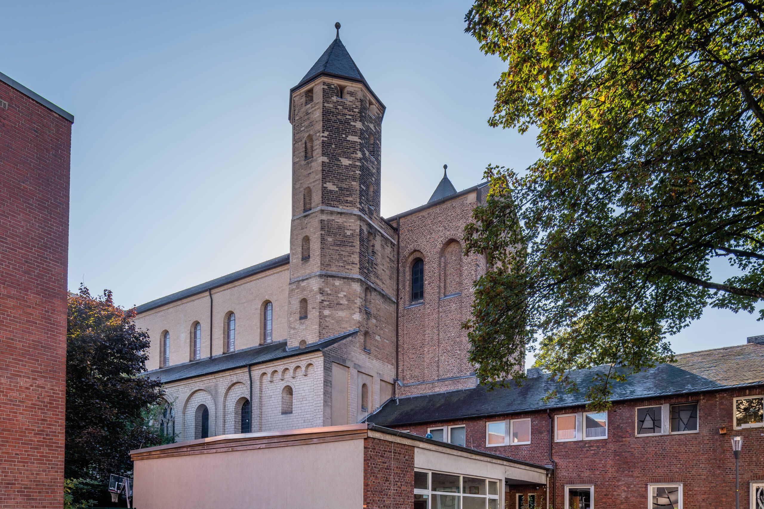 Die Kirche Sankt Maria im Kapitol in Köln, umgeben von Bäumen und angrenzenden Gebäuden, bei klarem Himmel.