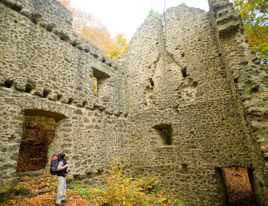 Ruine im Wald mit Herbstlaub und Wanderer. Die Bäume sind herbstlich gefärbt, und Laub bedeckt den Boden.