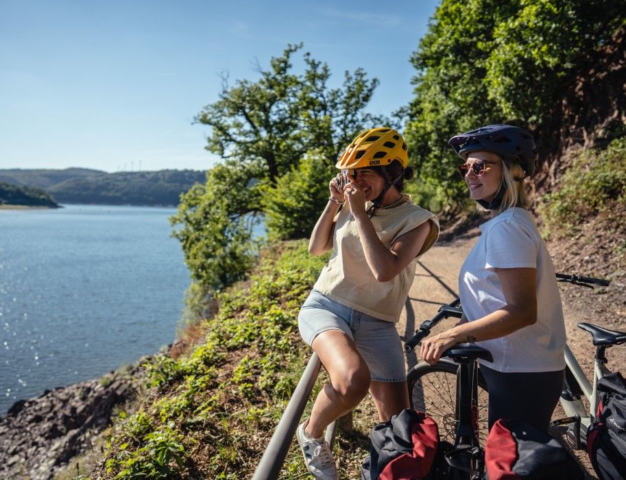 Zwei Frauen mit Fahrradhelmen stehen an einem See und machen Fotos.