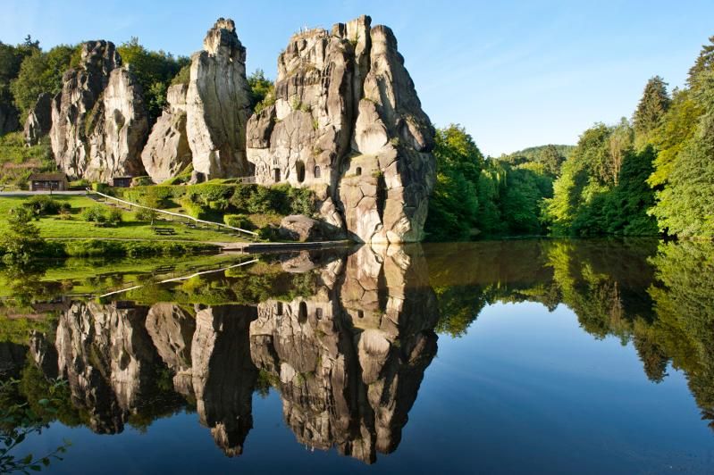 Die Externsteine spiegeln sich in einem ruhigen See, umgeben von grüner Vegetation und blauem Himmel.