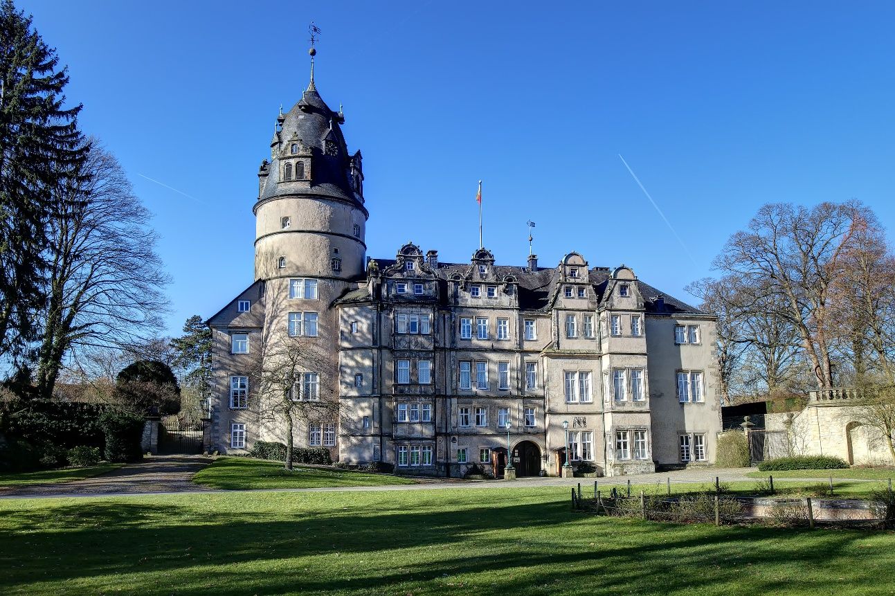 Fürstliches Residenzschloss Detmold mit rundem Turm und vielen Fenstern, umgeben von Bäumen und einem gepflegten Rasen. Der Himmel ist klar und blau.