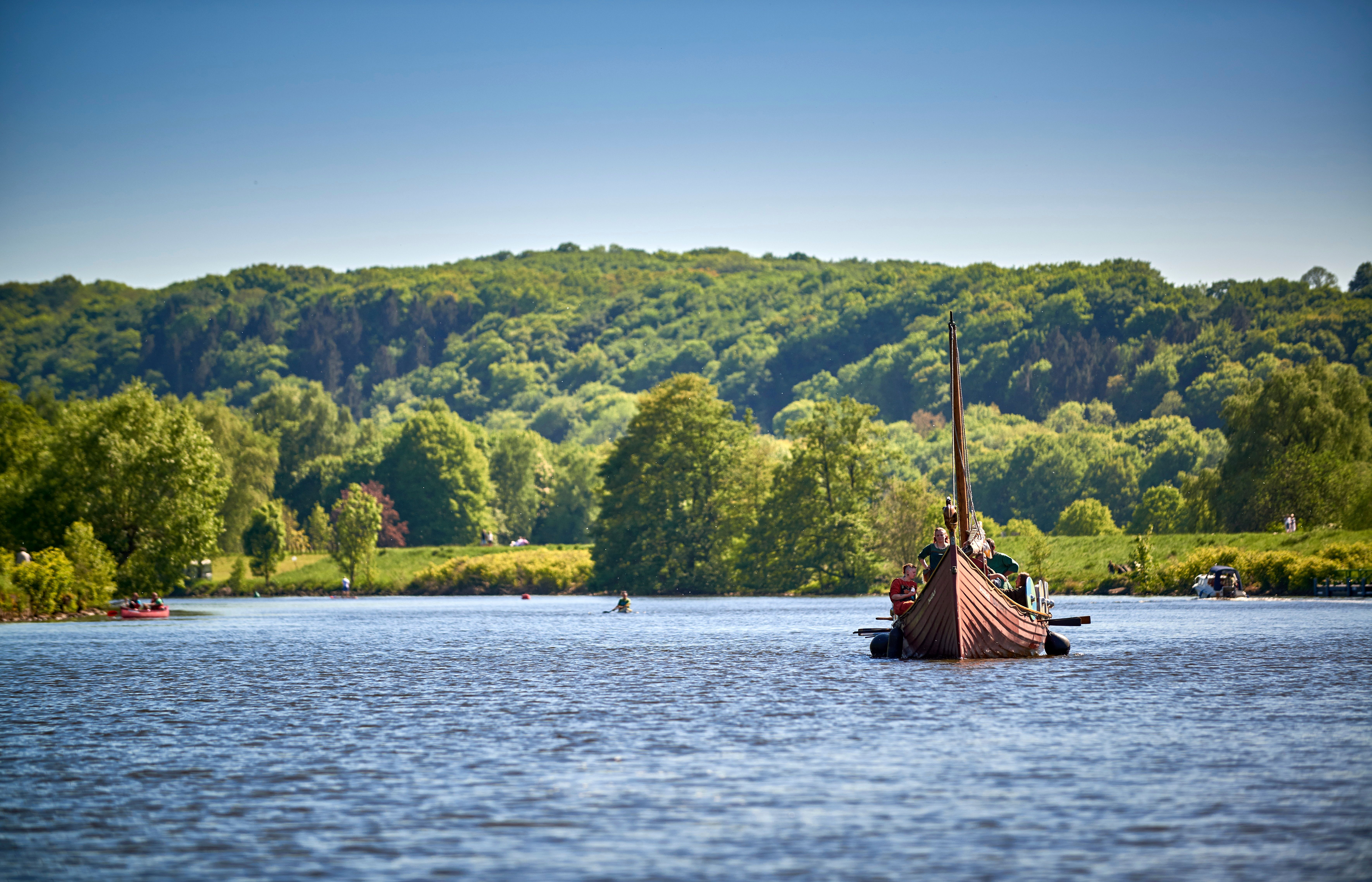 Ein Wikingerschiff fährt auf einem Fluss, umgeben von grüner Landschaft und bewaldeten Hügeln unter klarem Himmel.