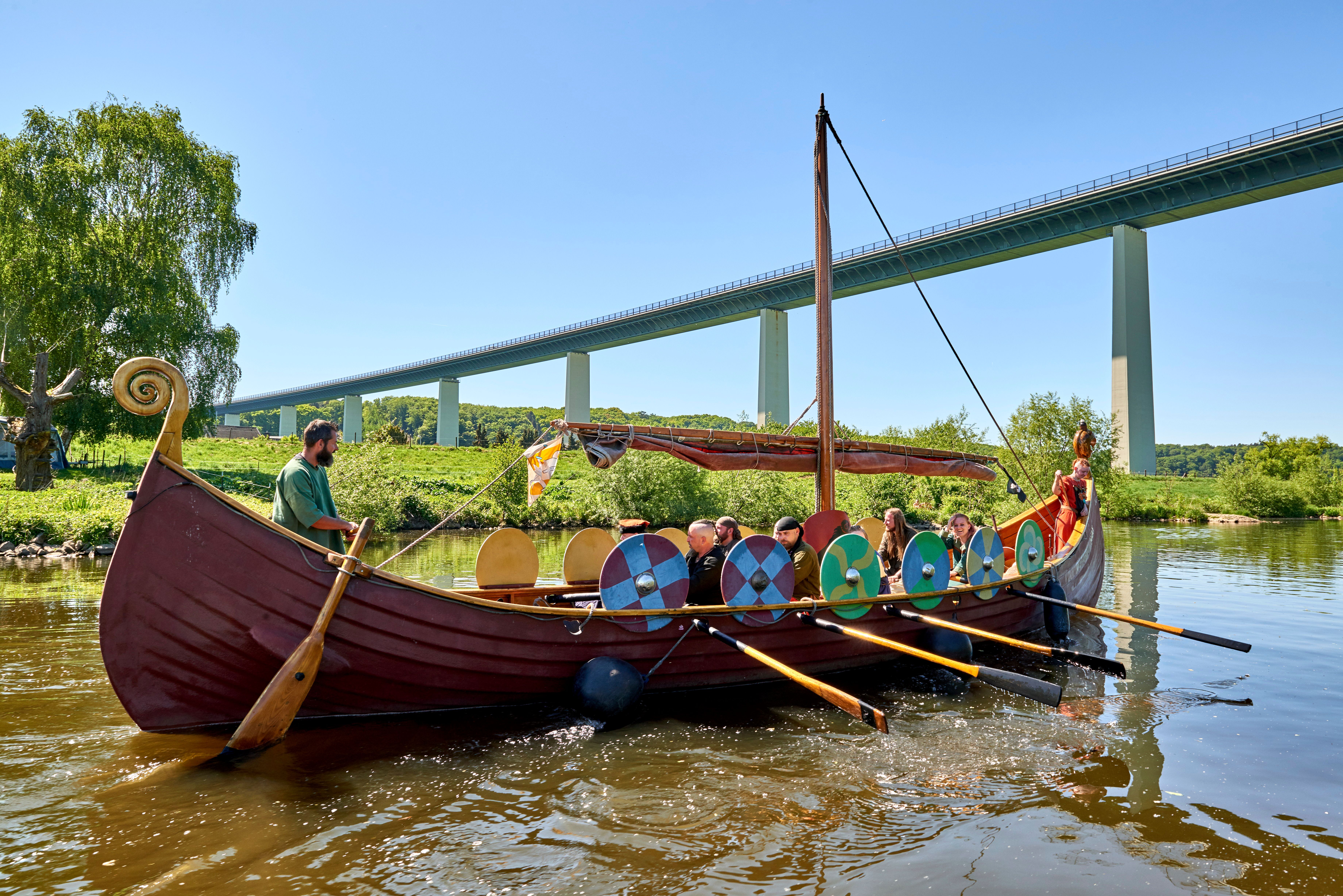 Ein Wikingerschiff mit Menschen auf einem Fluss, im Hintergrund eine Brücke und grüne Landschaft.