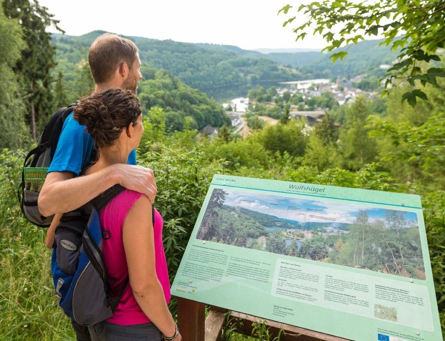 Ein Paar betrachtet eine Informationstafel mit der Aufschrift "Wolfshügel" in einer grünen Landschaft.