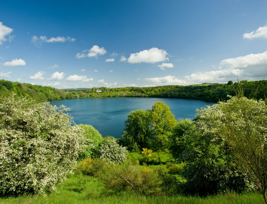 Das Weinfelder Maar in der Eifel ist umgeben von blühenden Bäumen und einem klaren blauen Himmel.