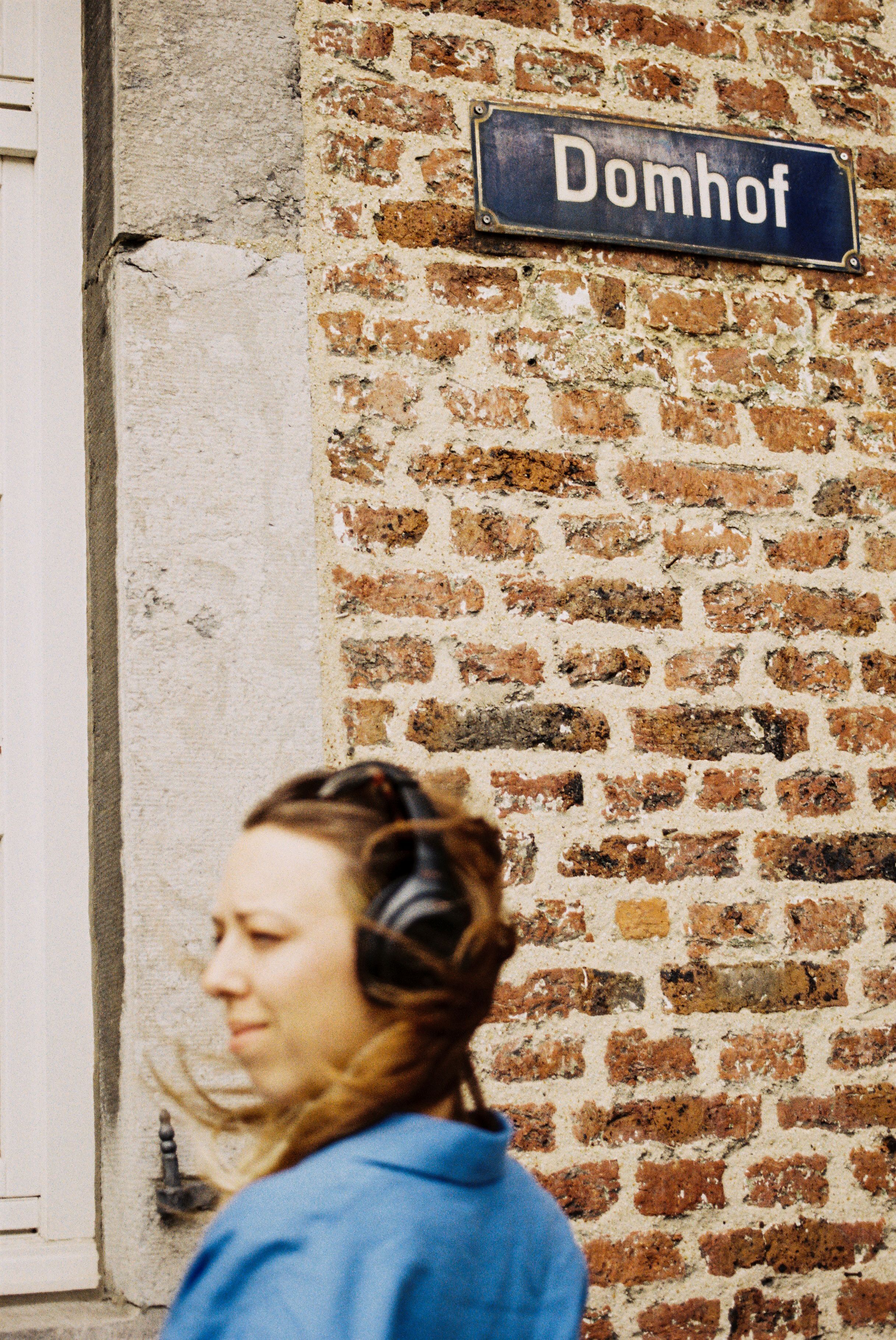 Frau mit Kopfhörern steht vor einer Ziegelsteinwand mit dem Straßenschild Domhof in Aachen.