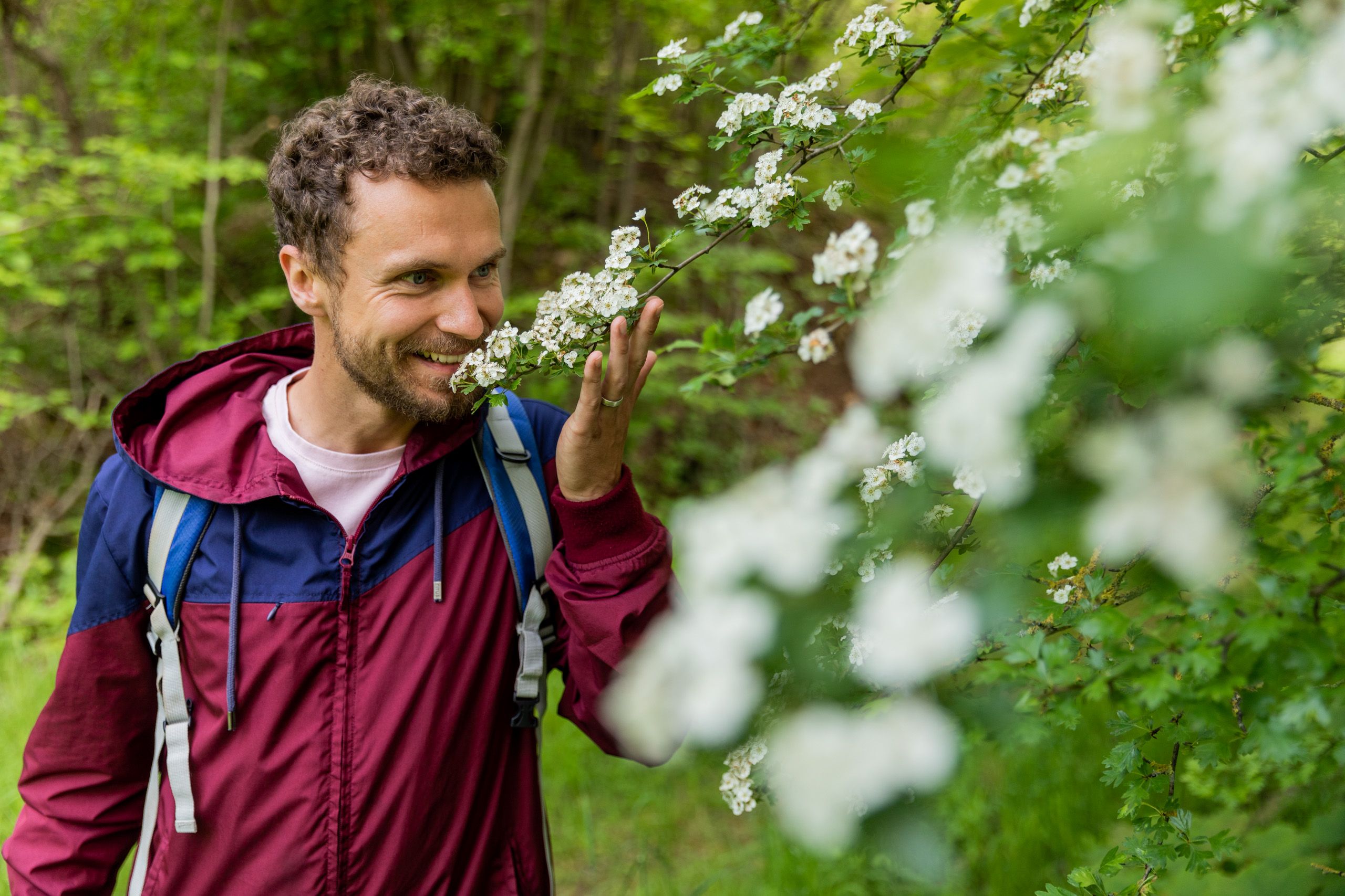Ein Mann steht in einer grünen Umgebung und lächelt, während er an weißen Blüten riecht.