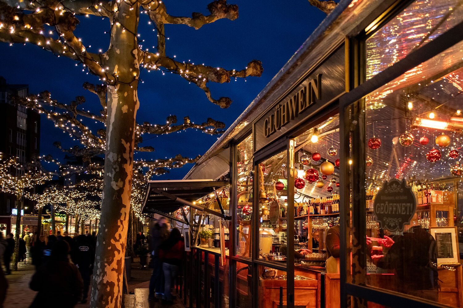 Ein festlich beleuchteter Weihnachtsmarkt mit einem Glühweinstand. Bäume sind mit Lichterketten geschmückt, und Menschen schlendern entlang der Promenade.