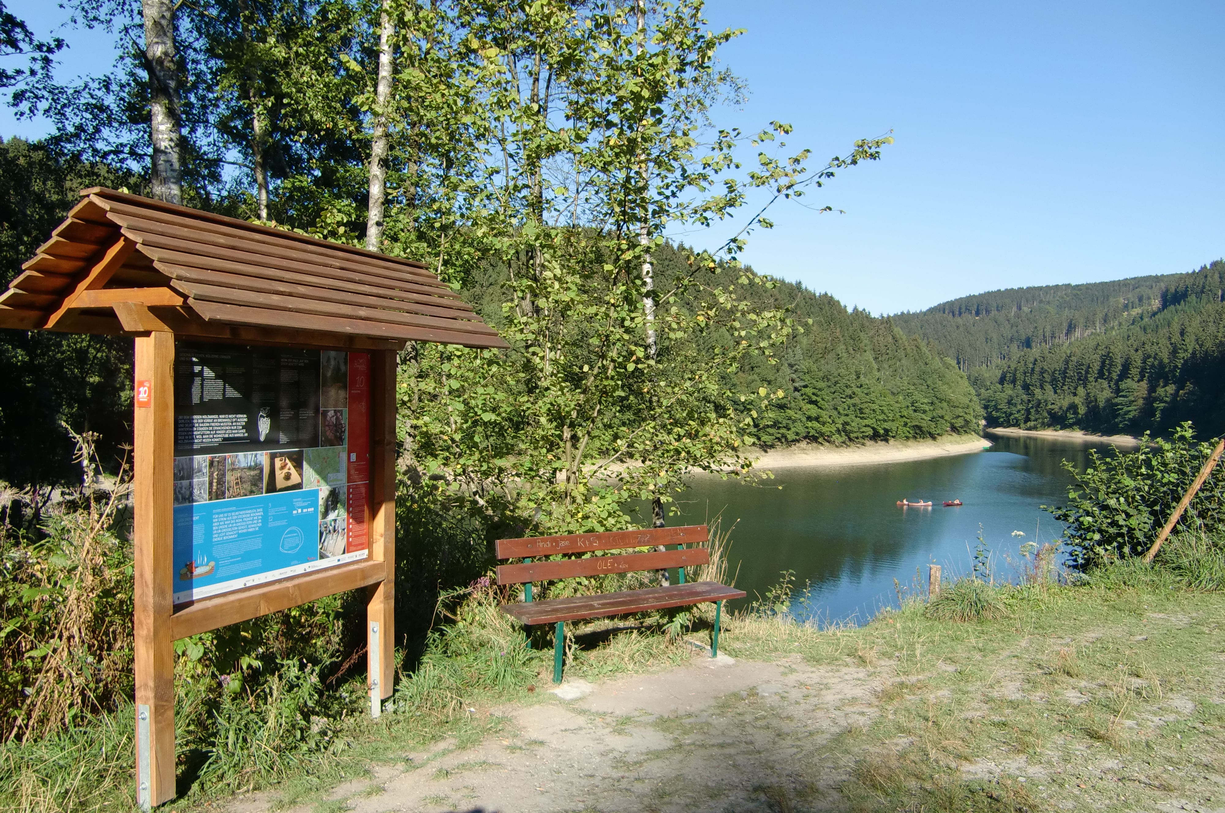 Infotafel und Bank am Ufer der Aggertalsperre mit bewaldeten Hügeln im Hintergrund.