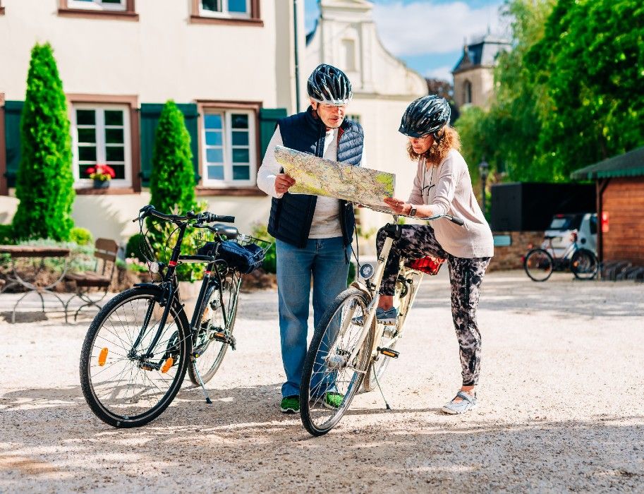 Zwei Radfahrer mit Helmen betrachten eine Karte vor einem Gebäude.