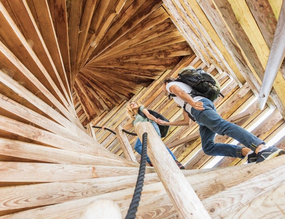 Zwei Personen steigen eine hölzerne Wendeltreppe hinauf, die spiralförmig nach oben führt.
