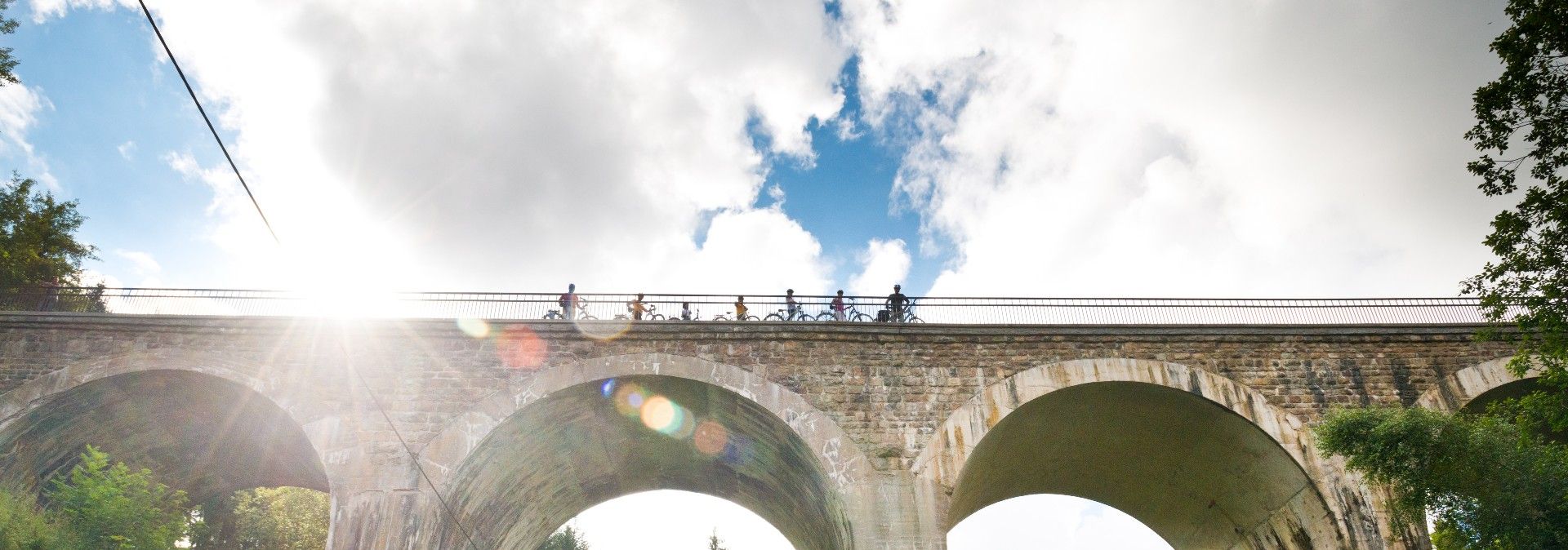 Fahrradfahrer überqueren den Vennbahn Viadukt, während die Sonne durch die Wolken scheint. Der Viadukt ist aus Stein mit großen Bögen.