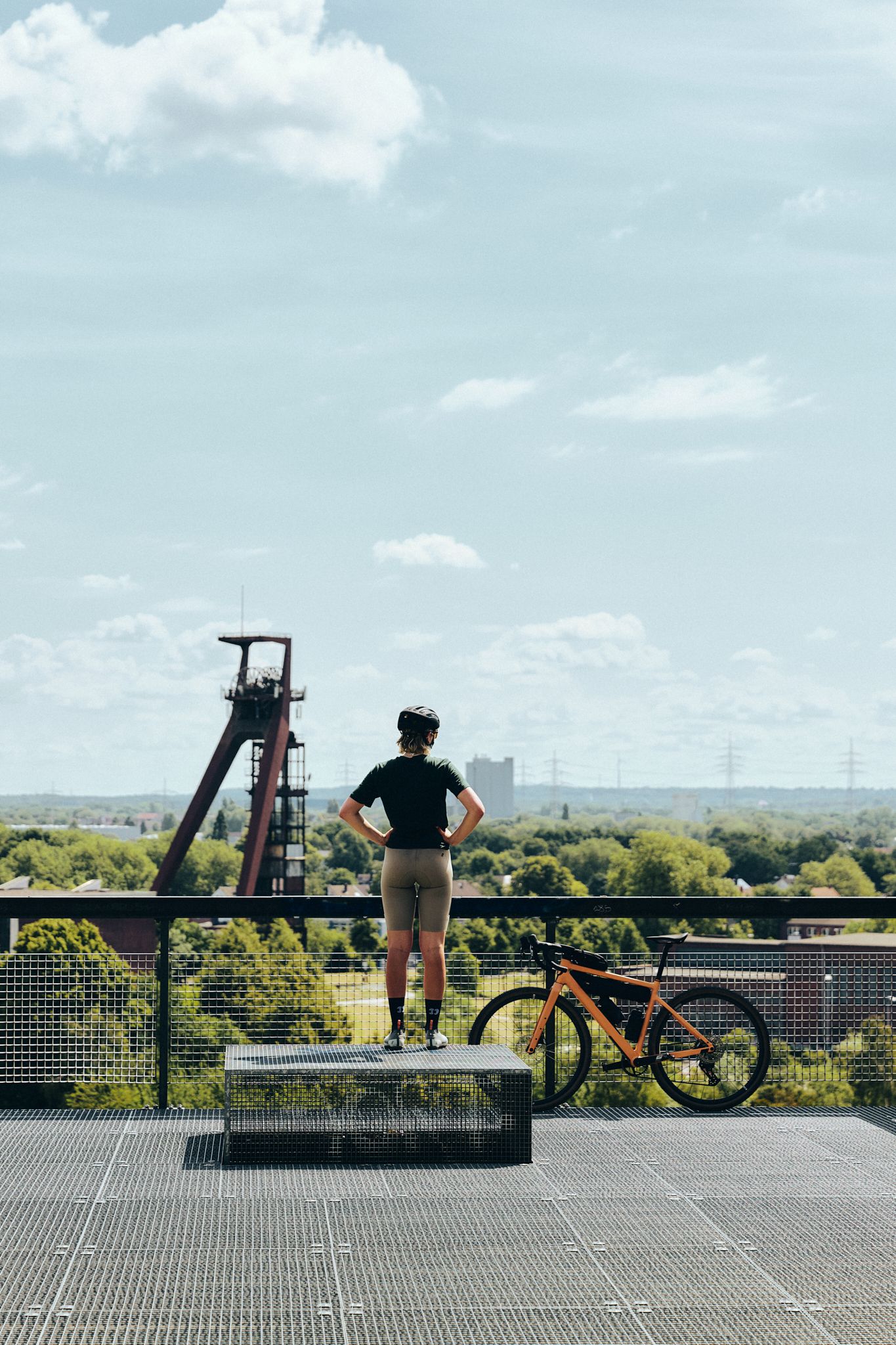 Eine Person mit Fahrradhelm steht auf einer Plattform und blickt auf eine grüne Landschaft mit einem Förderturm im Hintergrund.