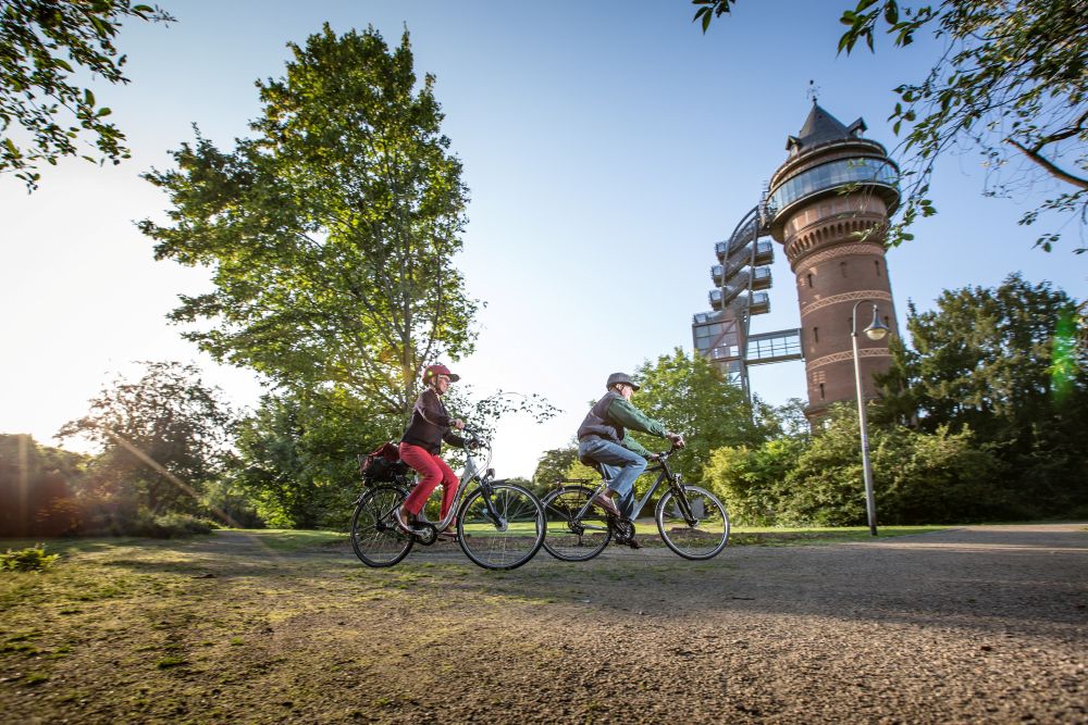 Zwei Radfahrer fahren auf dem RuhrtalRadweg, im Hintergrund ein Turm und Bäume.