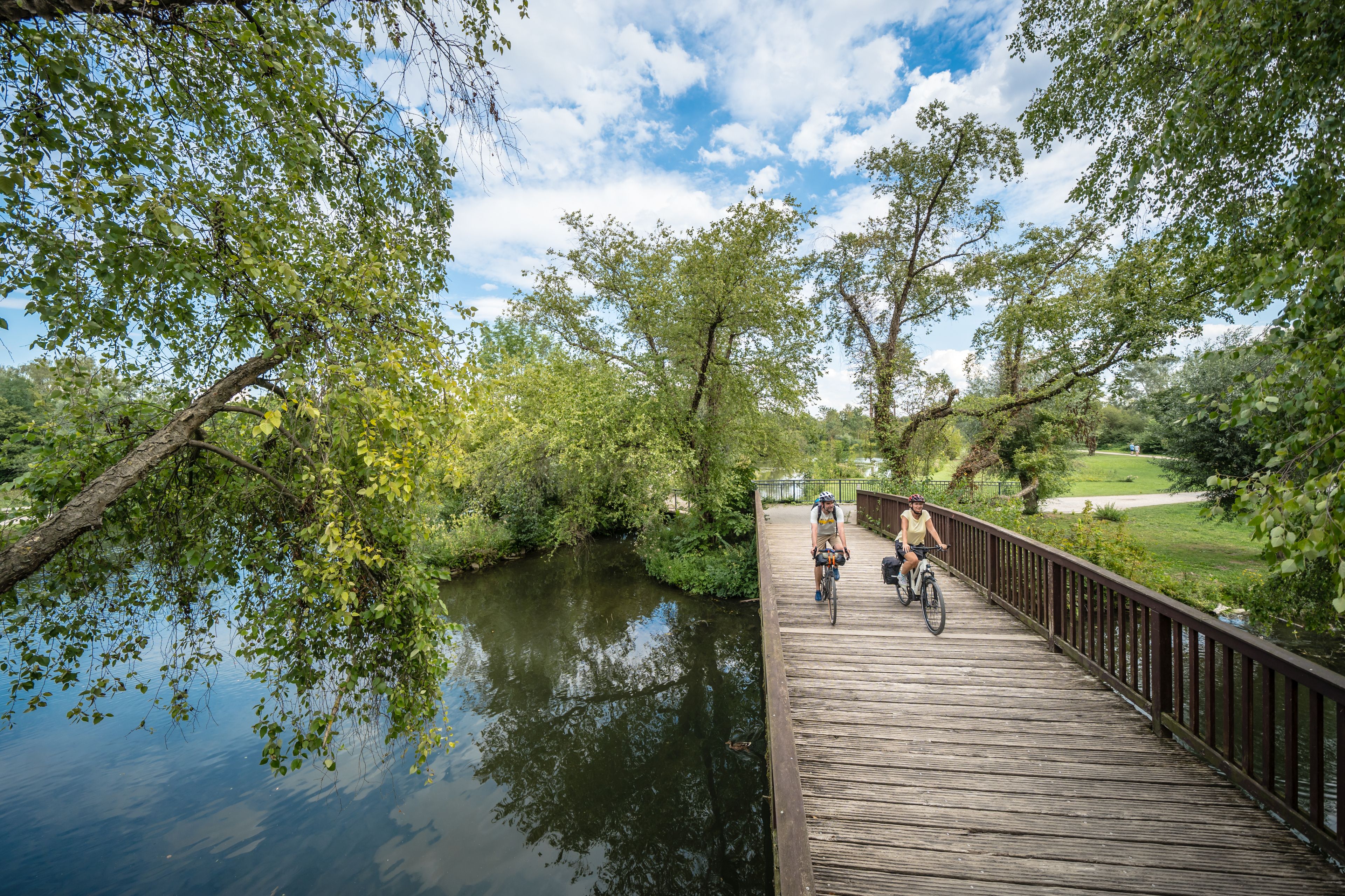 Zwei Radfahrer auf einer Holzbrücke über einem Fluss, umgeben von grünen Bäumen und blauem Himmel.