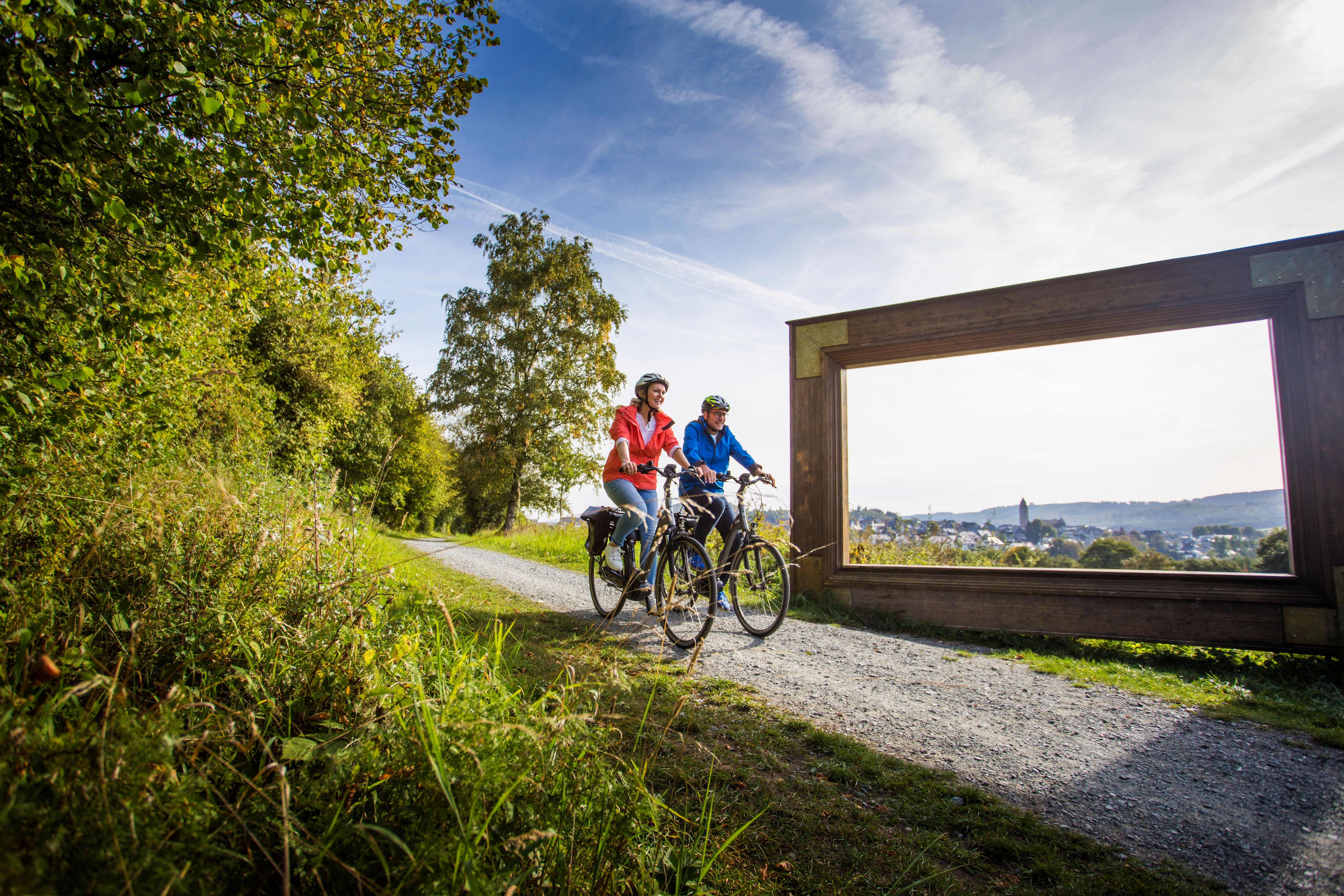 Zwei Radfahrer rollen entspannt über einen geschotterten Abschnitt des SauerlandRadrings. Am Wegesrand rahmt ein großes Holzfenster die weite Aussicht auf die Dächer des Ortes und die hügelige Landschaft im Hintergrund