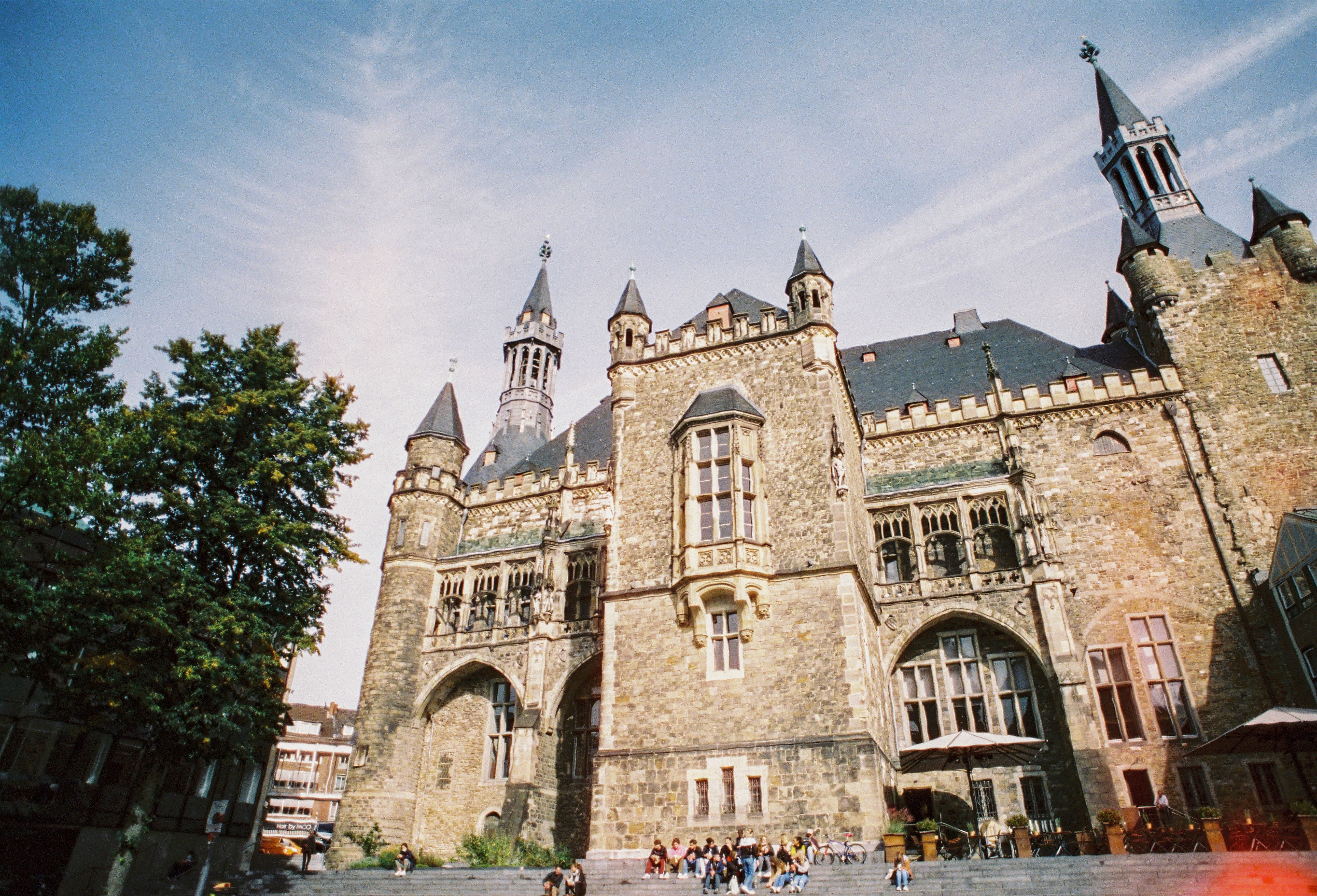 Das Aachener Rathaus mit seinen markanten Türmen und gotischen Elementen, umgeben von Bäumen und Menschen auf den Stufen davor.