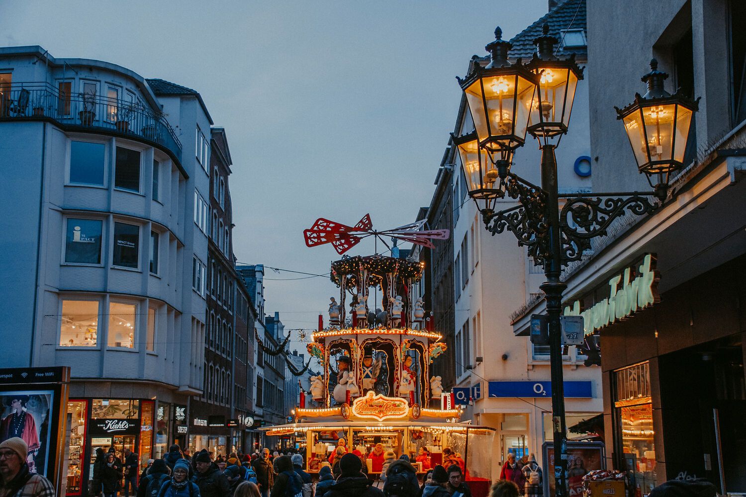 Altstadt-Markt in Düsseldorf mit beleuchteter Weihnachtskarussell-Pyramide in einer belebten Einkaufsstraße bei Dämmerung.