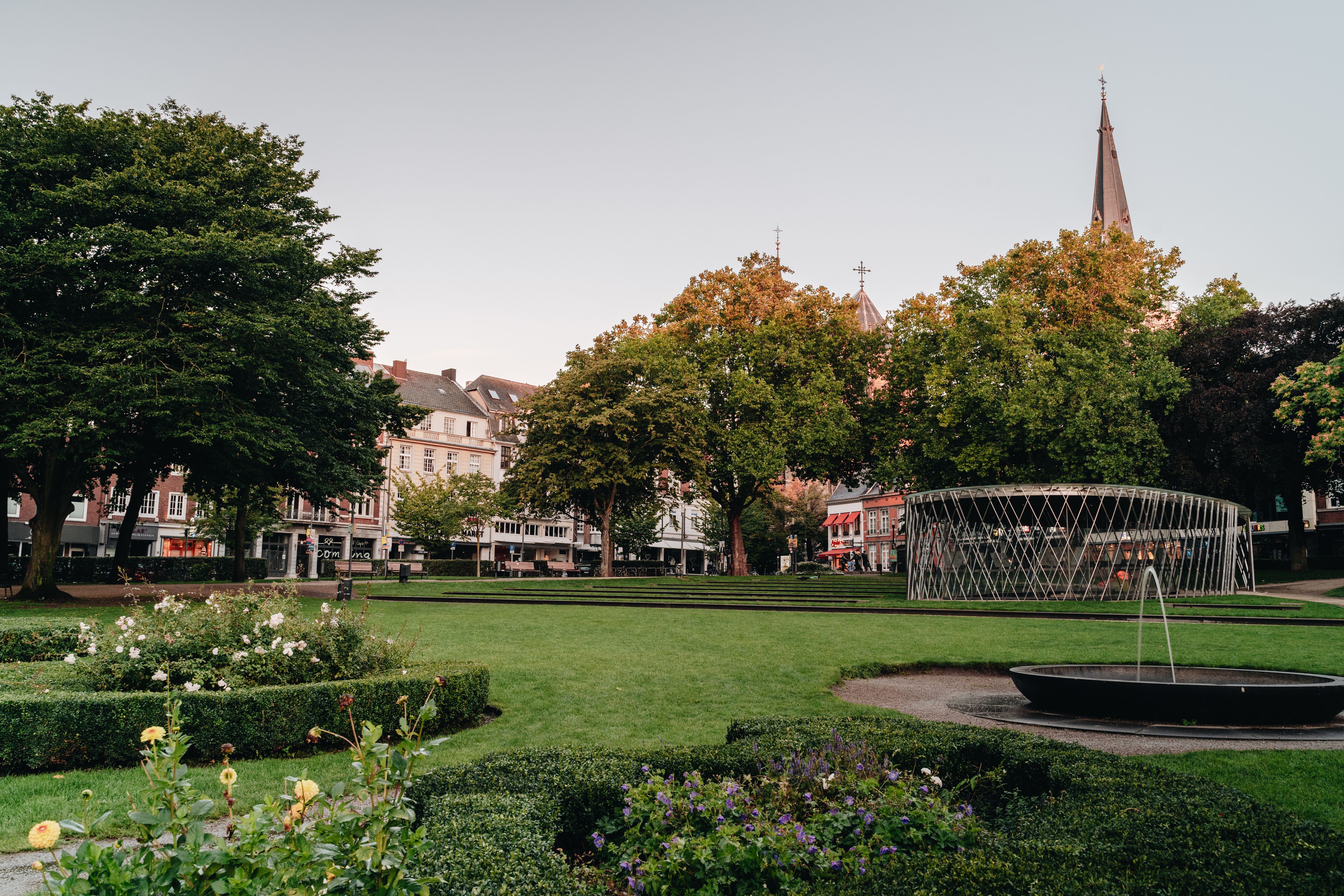 Der Elisengarten in Aachen zeigt eine grüne Parklandschaft mit einer archäologischen Vitrine und einem kleinen Springbrunnen.