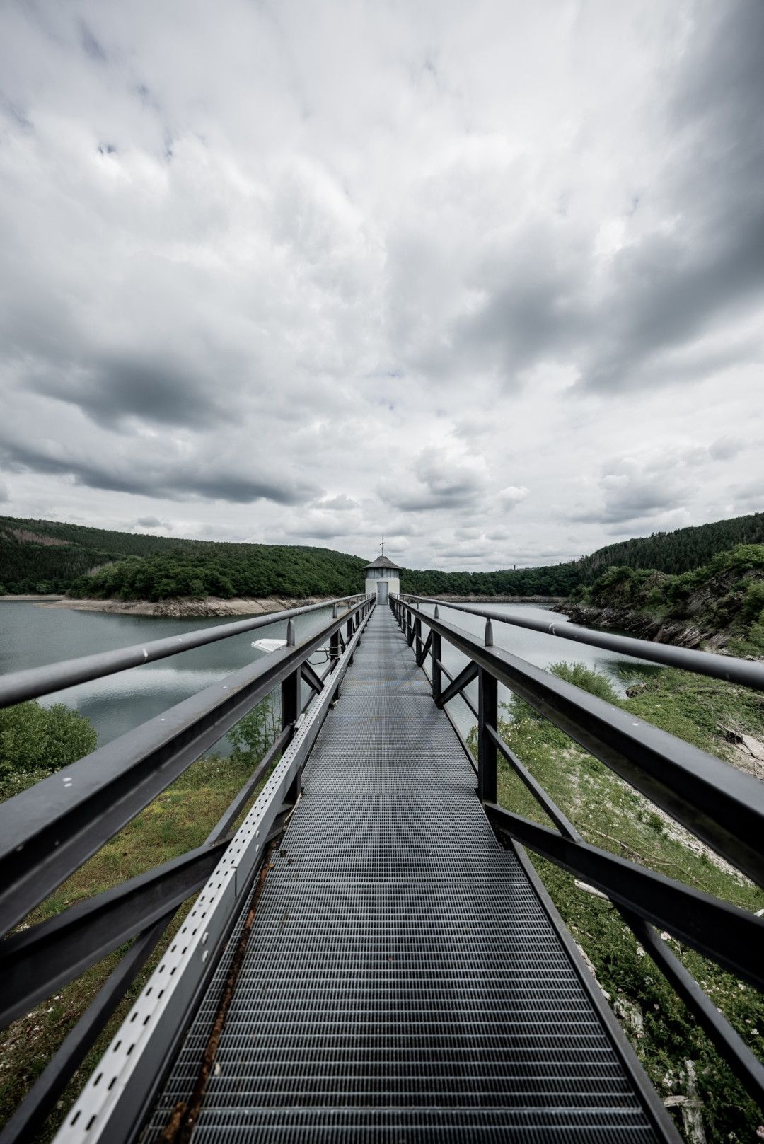 Ein langer Steg führt über die Urfttalsperre in der Eifel, umgeben von Wasser und bewaldeten Hügeln unter einem bewölkten Himmel.