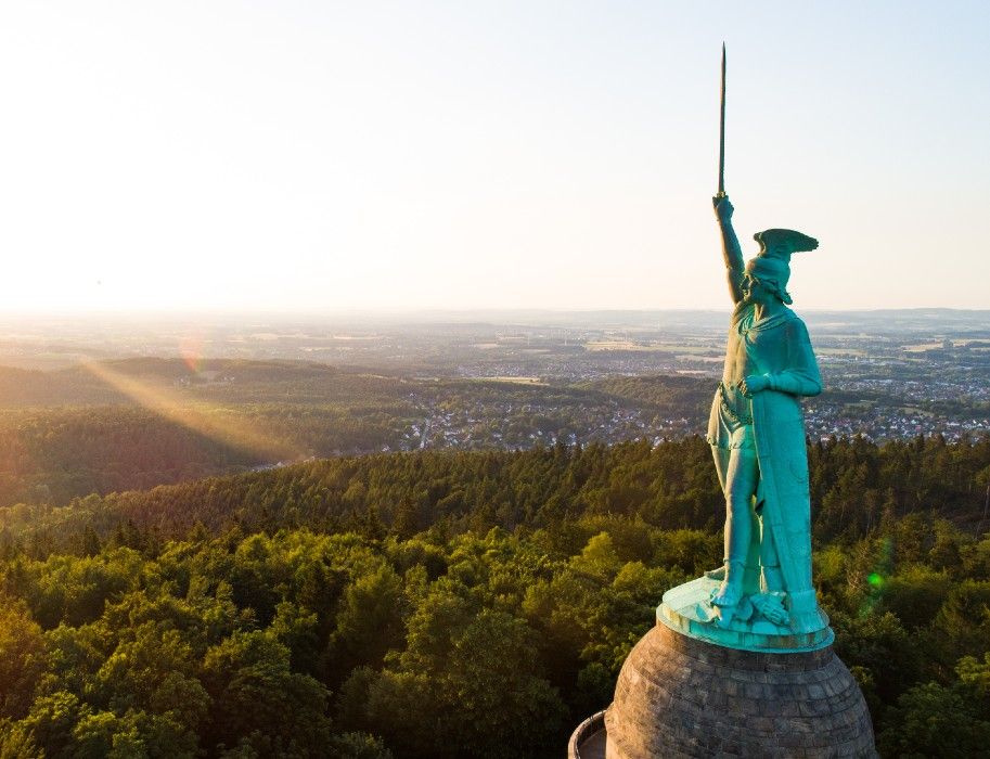 Hermannsdenkmal im Teutoburger Wald bei Sonnenuntergang, mit Blick auf bewaldete Hügel und eine Stadt im Hintergrund.