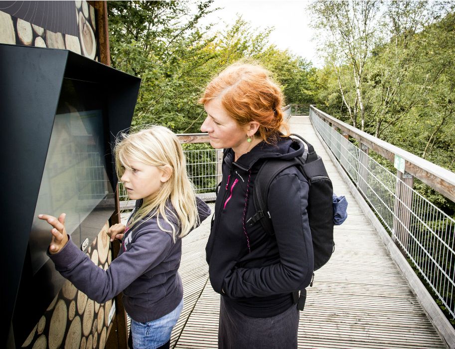 Eine Frau und ein Kind interagieren mit einem Bildschirm auf einem Holzsteg im Wald.