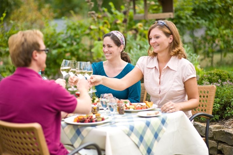 Drei Personen stoßen mit Weingläsern an einem Tisch im Freien an.  Der Tisch ist mit Speisen gedeckt, die Umgebung ist grün.