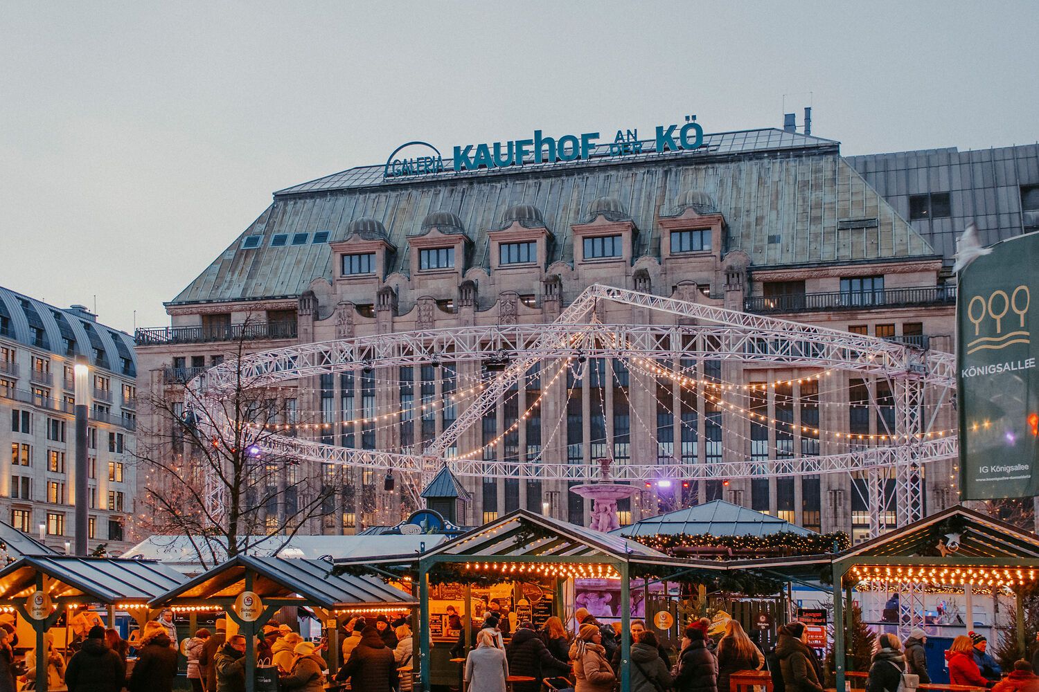 Wintermarkt mit beleuchteten Ständen und Menschen. Im Hintergrund das Gebäude "Galeria Kaufhof an der Kö". Lichterketten schmücken die Szene.