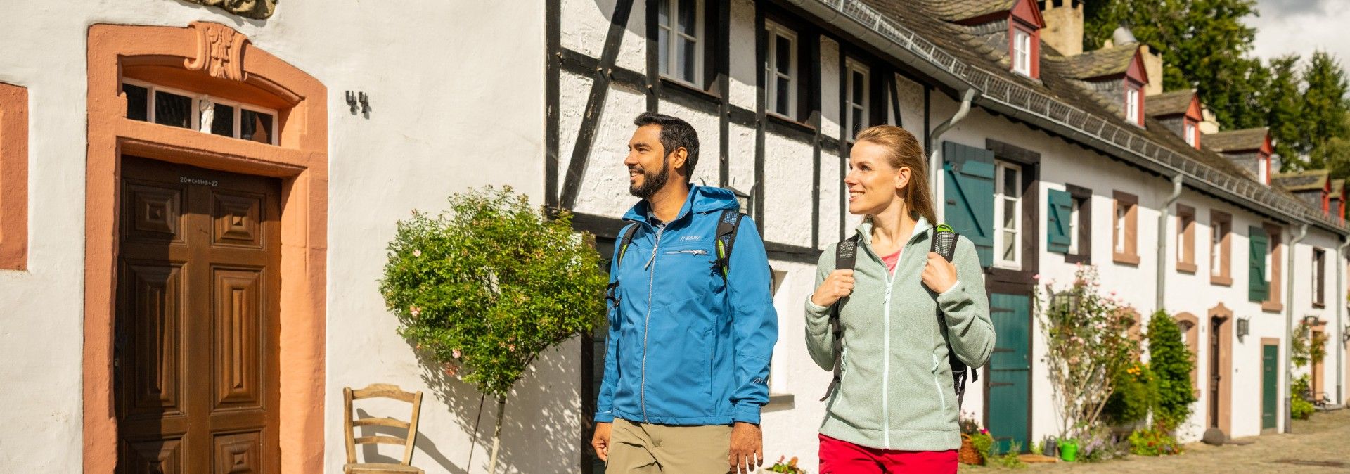 Zwei Personen mit Rucksäcken spazieren an einer Reihe von Fachwerkhäusern vorbei. Die Sonne scheint, und die Umgebung wirkt idyllisch.