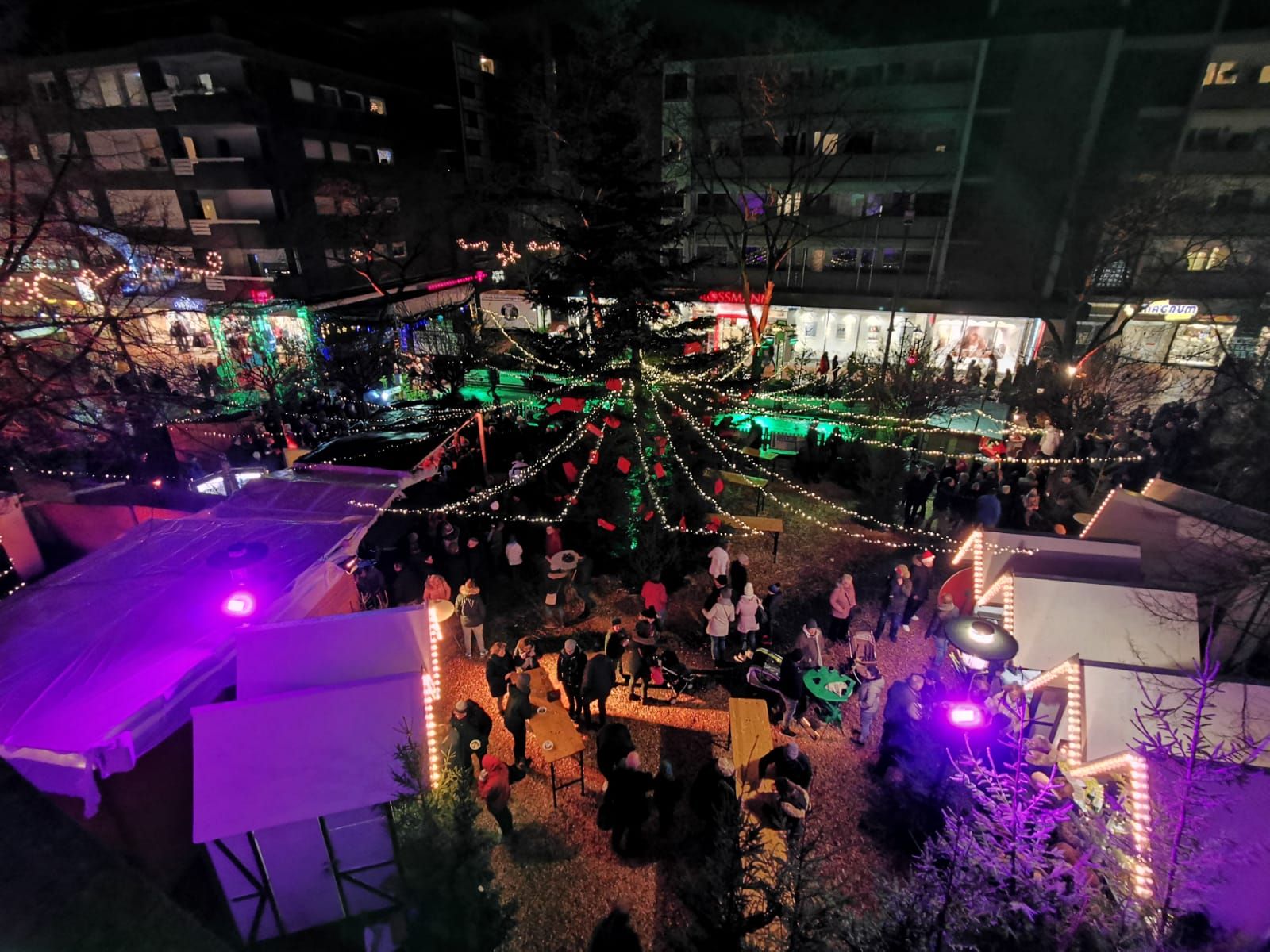 Luftaufnahme des Weihnachtsmarkts in Gronau bei Nacht. Menschen schlendern zwischen festlich beleuchteten Ständen und einem großen Weihnachtsbaum.