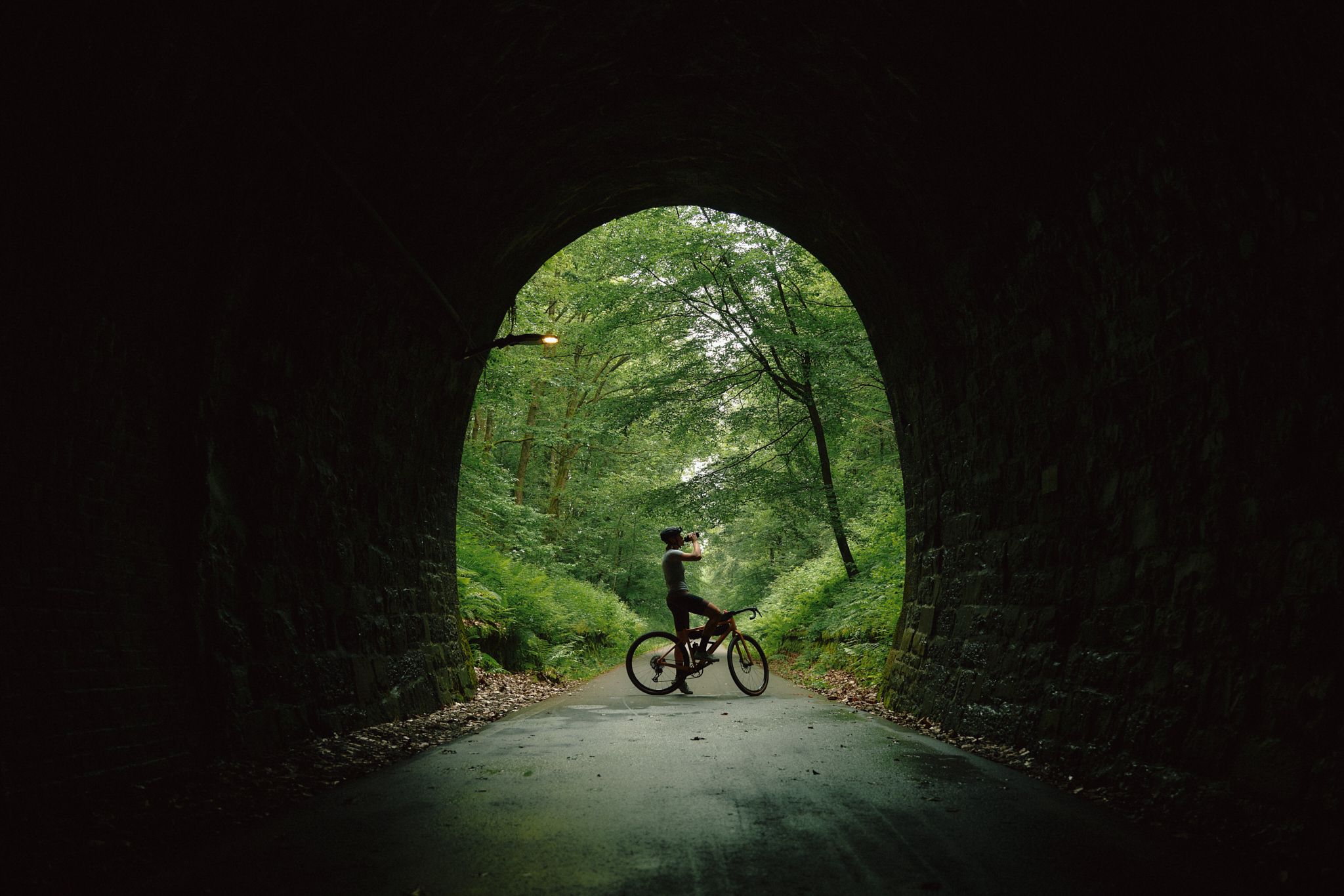 Radfahrende trinkt aus einer Flasche in einem Tunnel mit Blick auf einen grünen Wald.