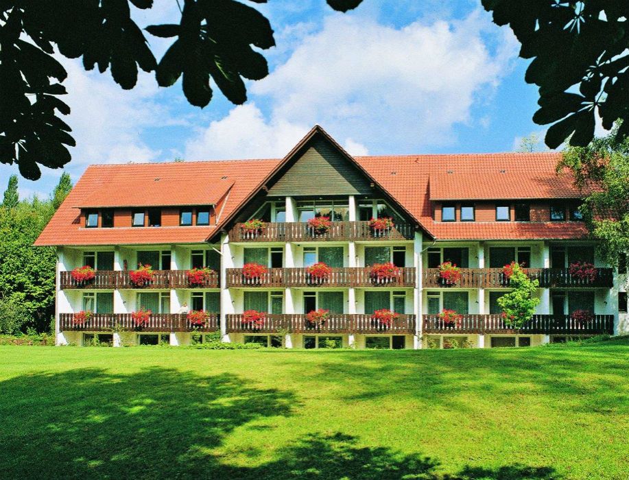 Ein dreistöckiges Gebäude mit roten Dachziegeln und Balkonen, die mit roten Blumen geschmückt sind, umgeben von einer grünen Wiese und Bäumen.