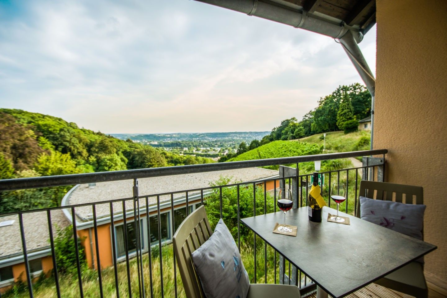 Balkon mit Tisch, zwei Weingläsern und Flasche, Blick auf grüne Landschaft und Stadt in der Ferne.