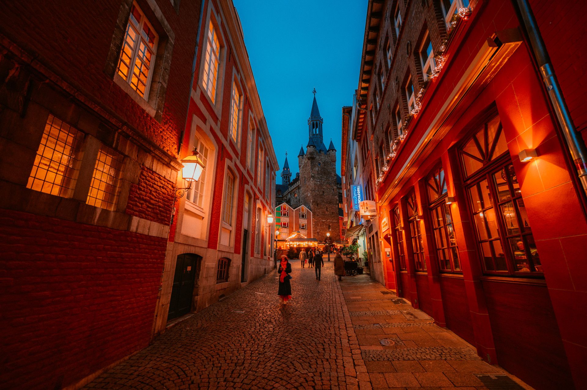 Eine beleuchtete Gasse in Aachen mit Kopfsteinpflaster und historischen Gebäuden, im Hintergrund der Aachener Dom.