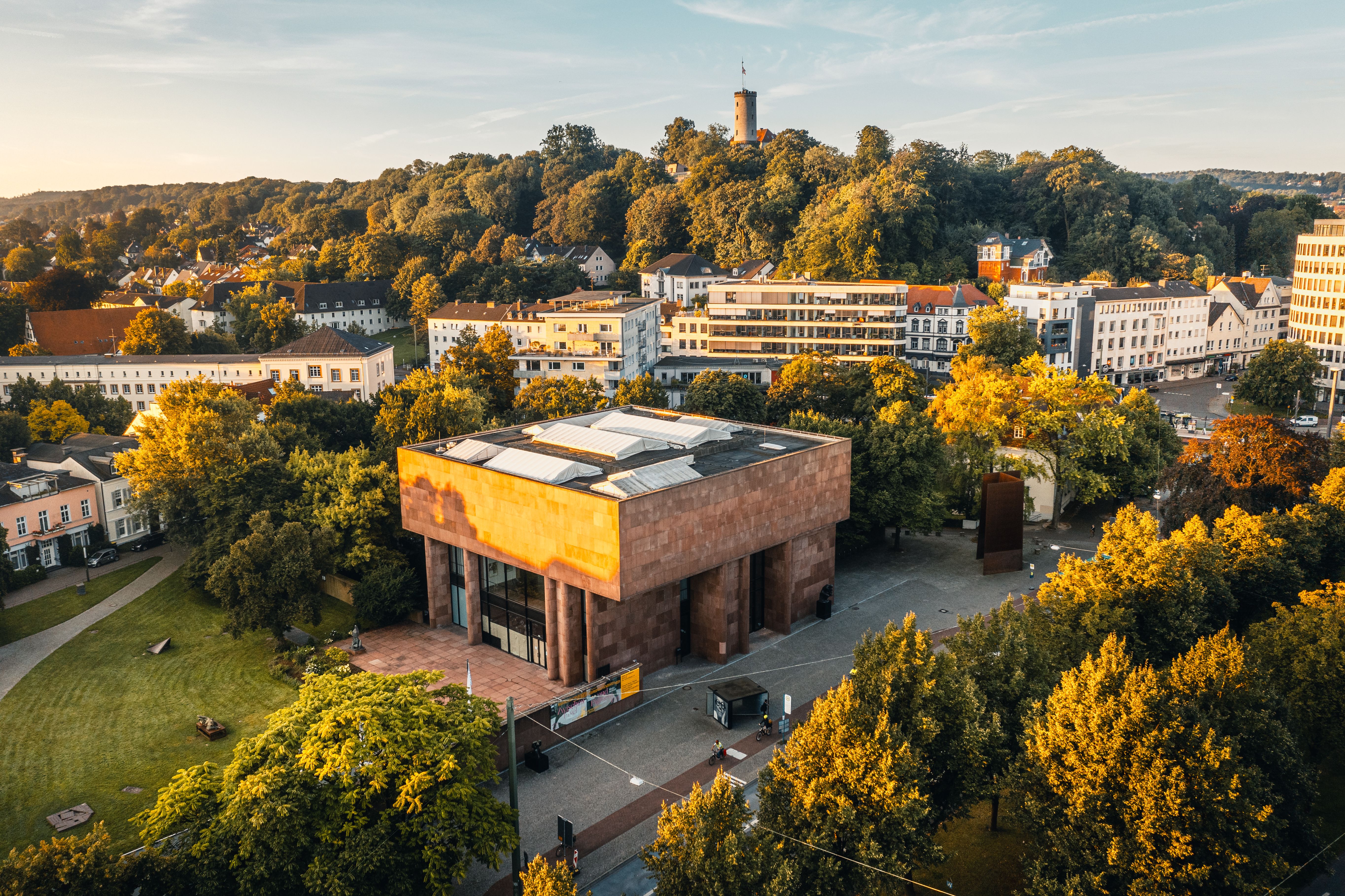 Die Kunsthalle Bielefeld im Vordergrund, umgeben von Bäumen und Gebäuden, mit der Sparrenburg auf einem bewaldeten Hügel im Hintergrund.
