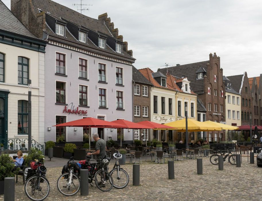 Historische Gebäude mit Cafés und bunten Sonnenschirmen. Fahrräder und Menschen auf einem gepflasterten Platz