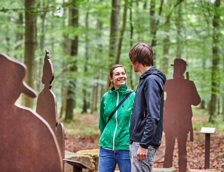 Zwei Personen stehen lächelnd im Wald neben Metallfiguren.