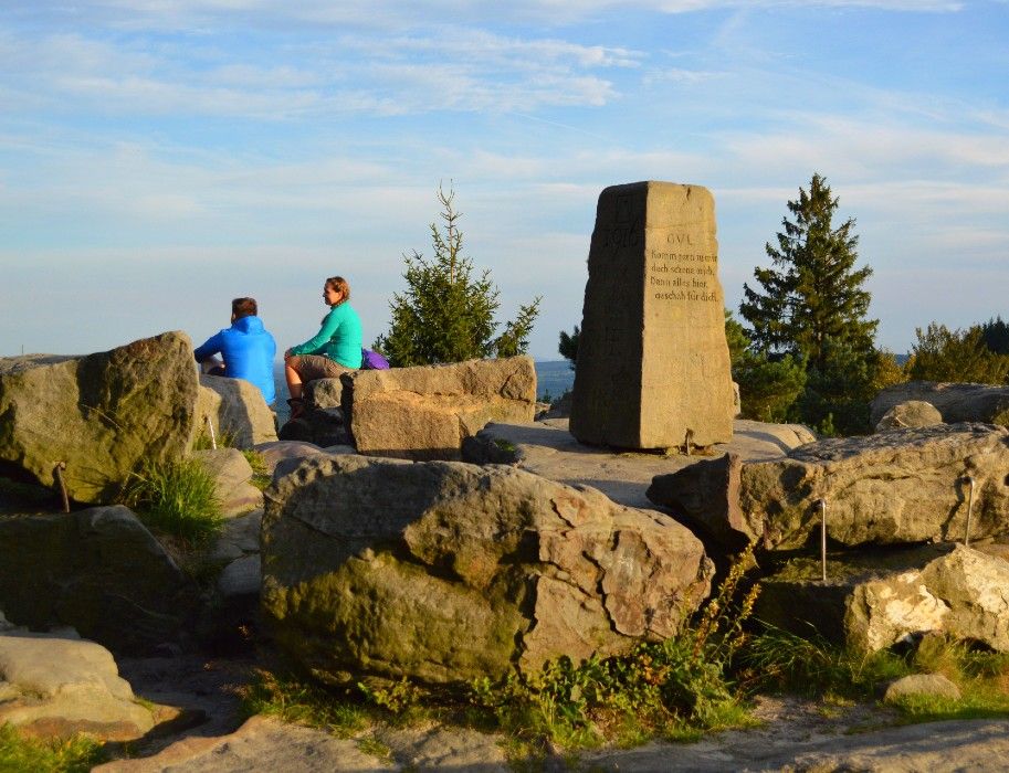 Zwei Personen sitzen auf Felsen am Lippischen Velmerstot. Ein Gedenkstein steht in der Mitte, umgeben von Bäumen und blauem Himmel.